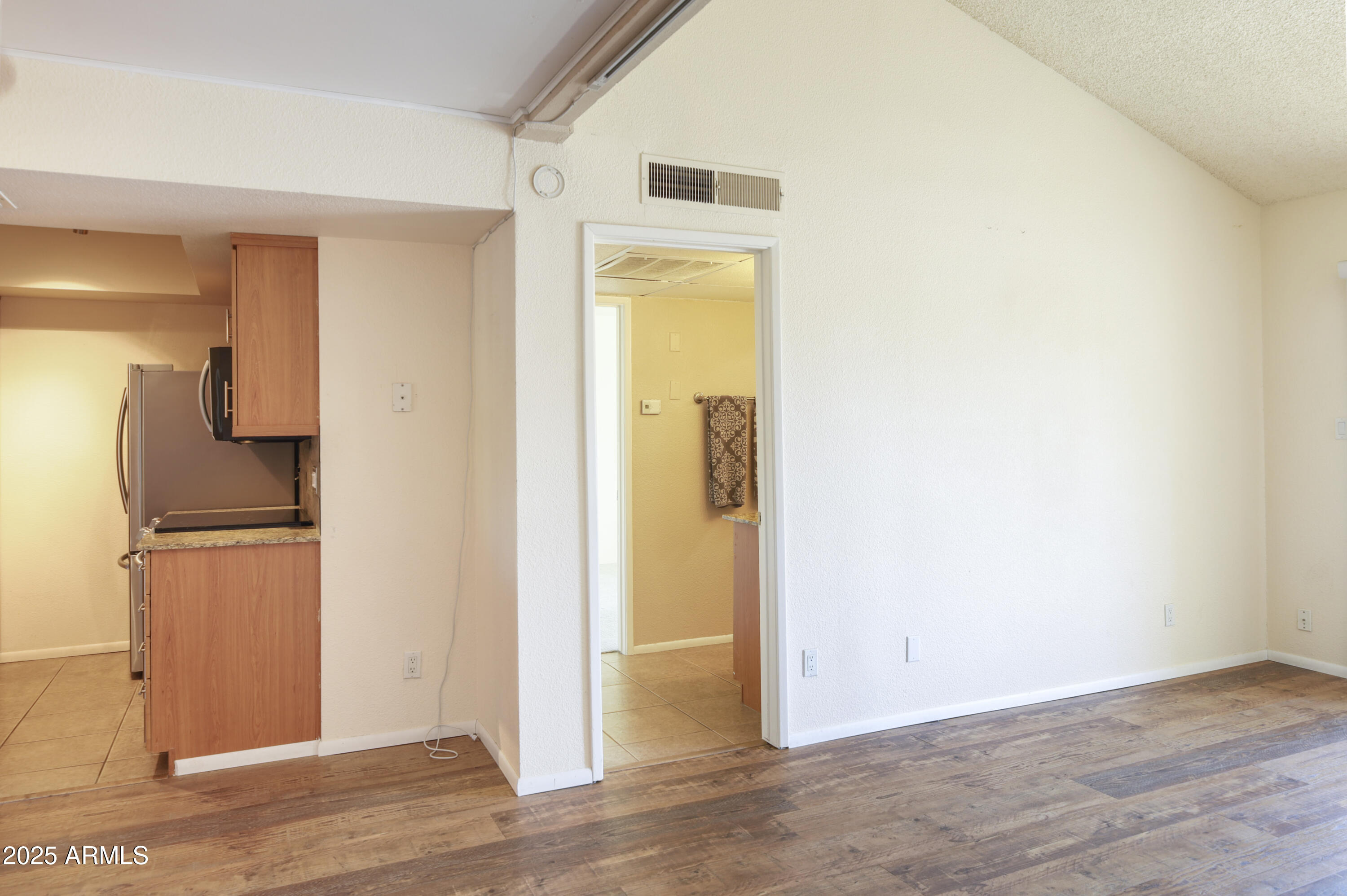 Vacant living area with wood-look floors, vaulted ceiling, and adjacent kitchen with wood cabinets and stainless refrigerator.