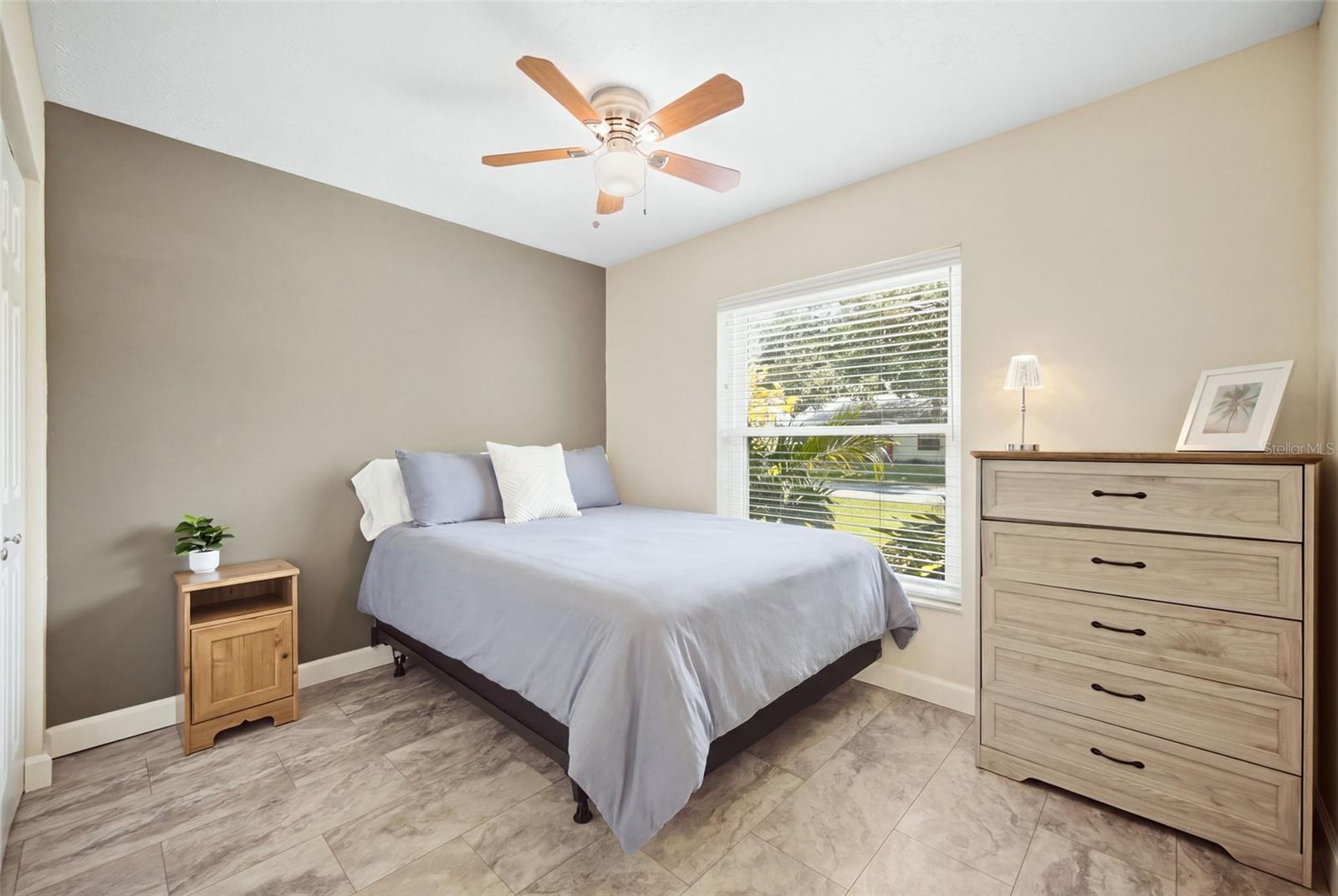 Bedroom with a gray-covered bed, wood furniture, ceiling fan, and a large window with blinds.