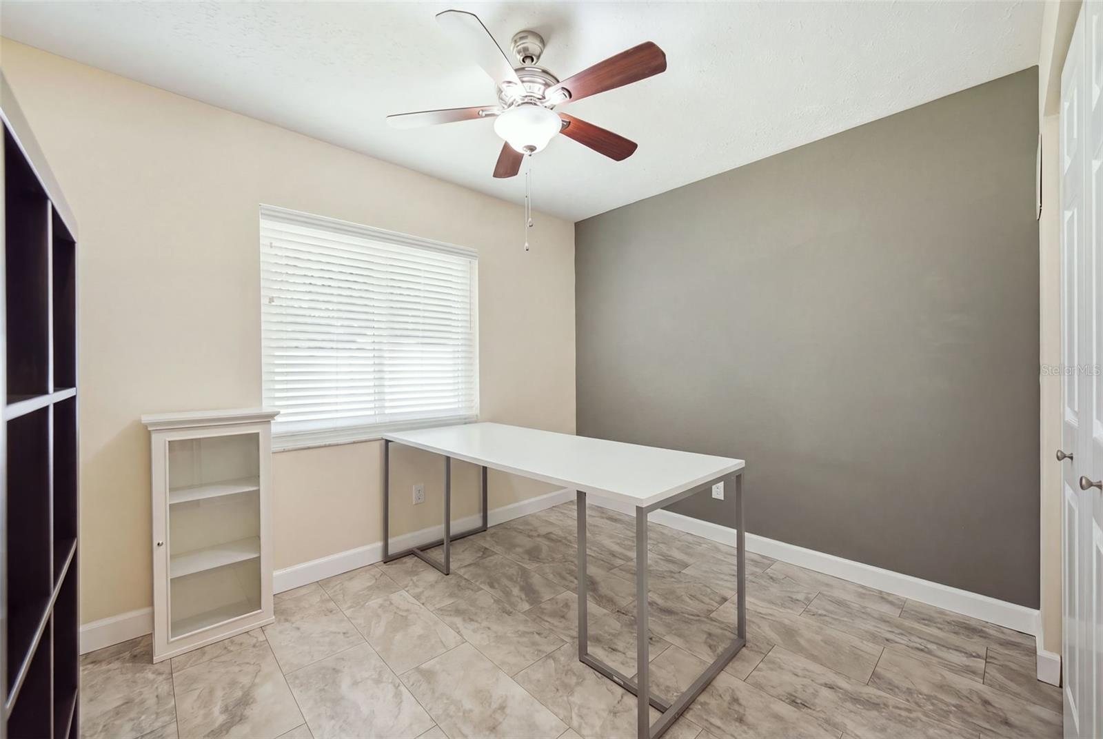 Home office with a white desk, gray accent wall, tiled floor, and window with blinds.