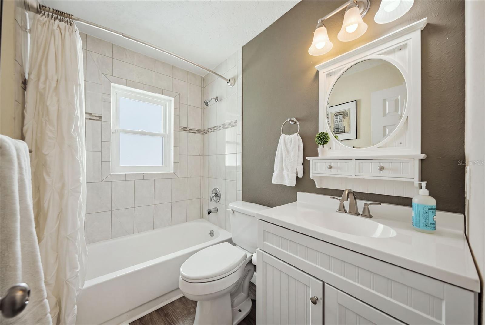 Bathroom with a tiled tub and shower, white vanity, and round mirror.