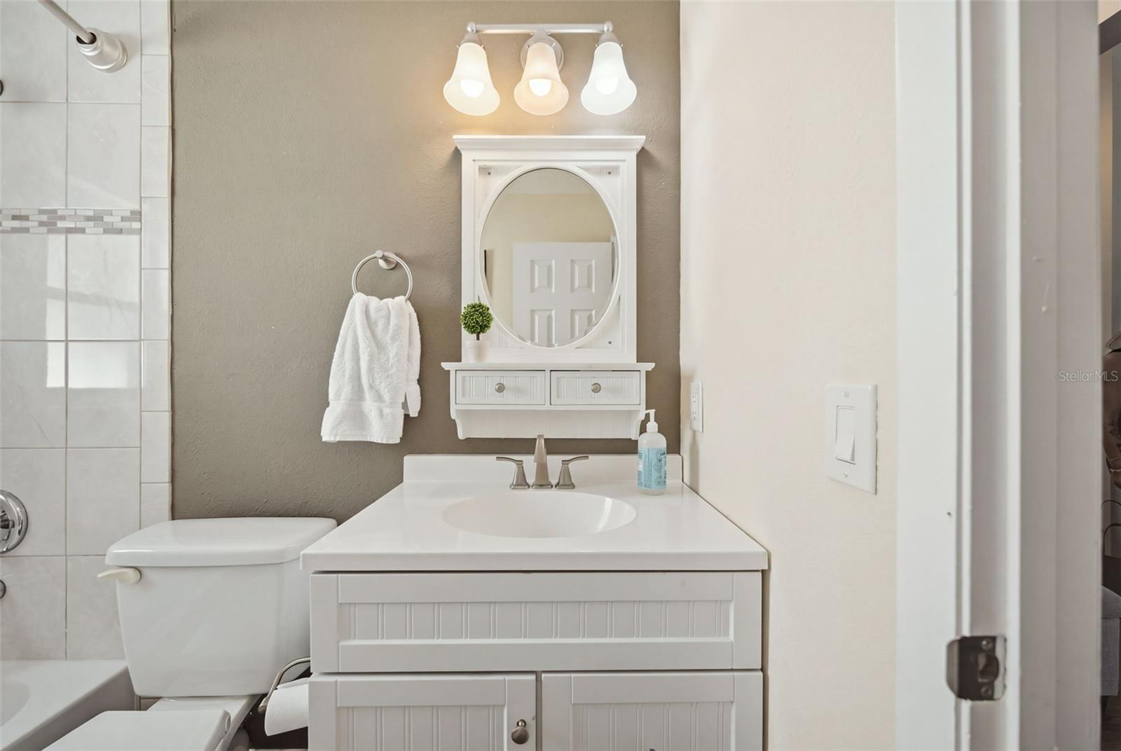 Bathroom vanity with an oval mirror beside a toilet and tiled tub surround.