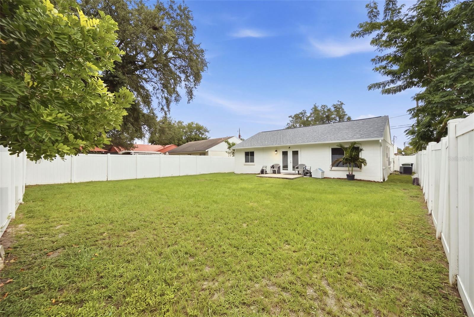 Fenced grassy backyard with a white house, small patio, and potted palm.