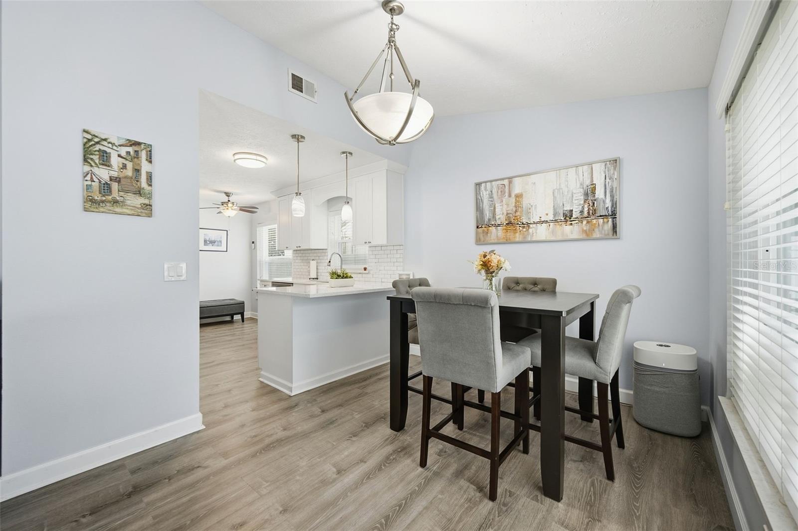 Dining area with gray chairs beside a bright white kitchen and wood-look flooring.