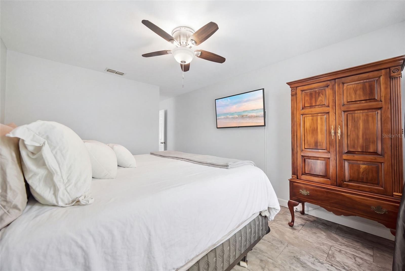 Bedroom with a white bed, carved wooden armoire, and ceiling fan.