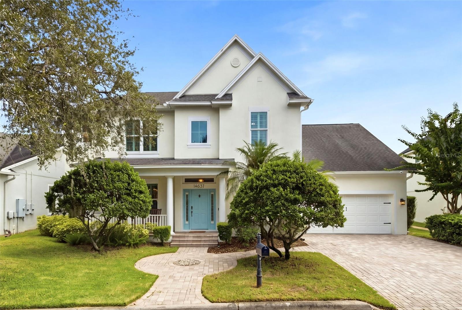 Two-story white stucco house with a blue front door, steep gables, landscaped lawn, and curved paved walkway.