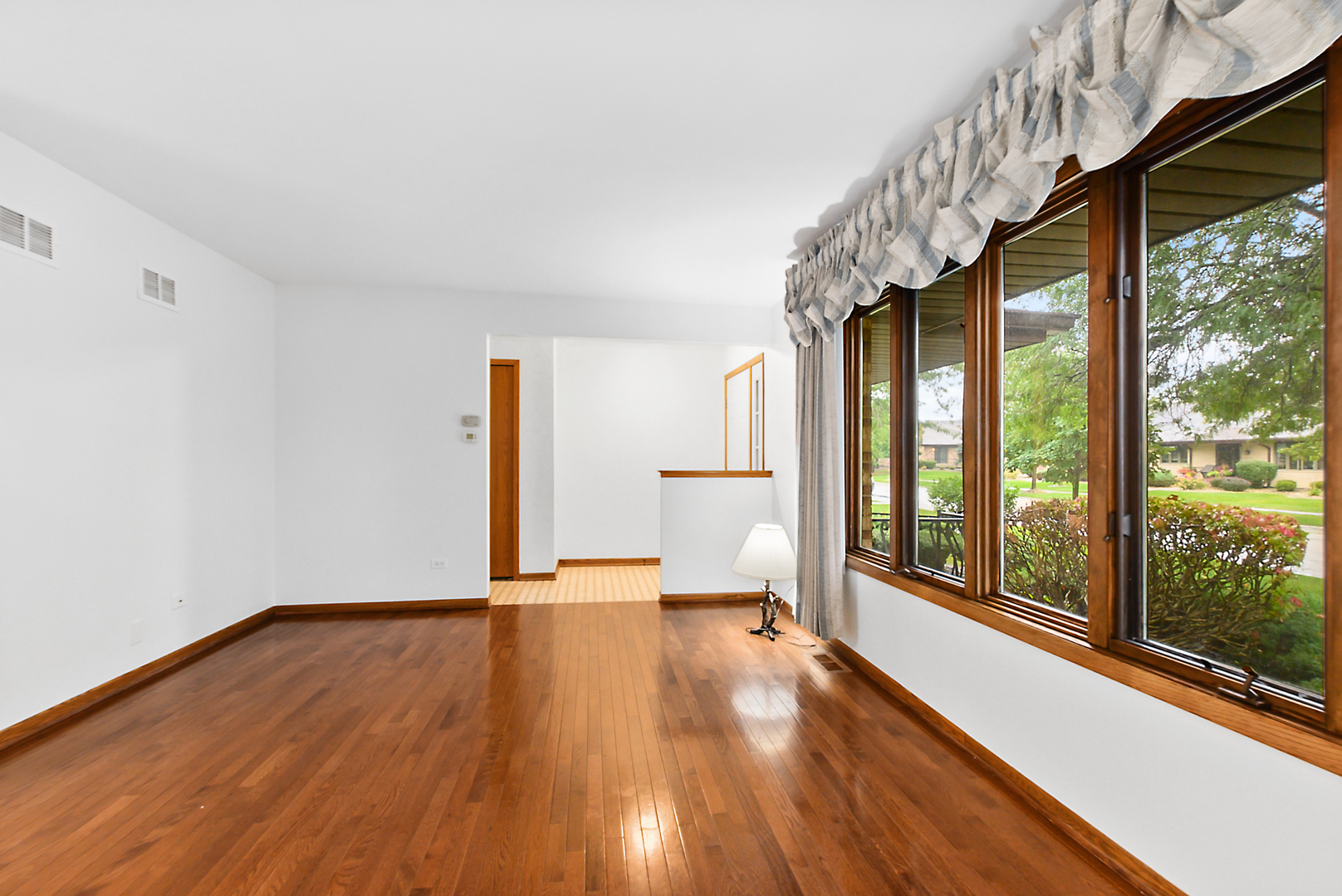 Empty living room with glossy hardwood floors and large wood-framed windows overlooking greenery.