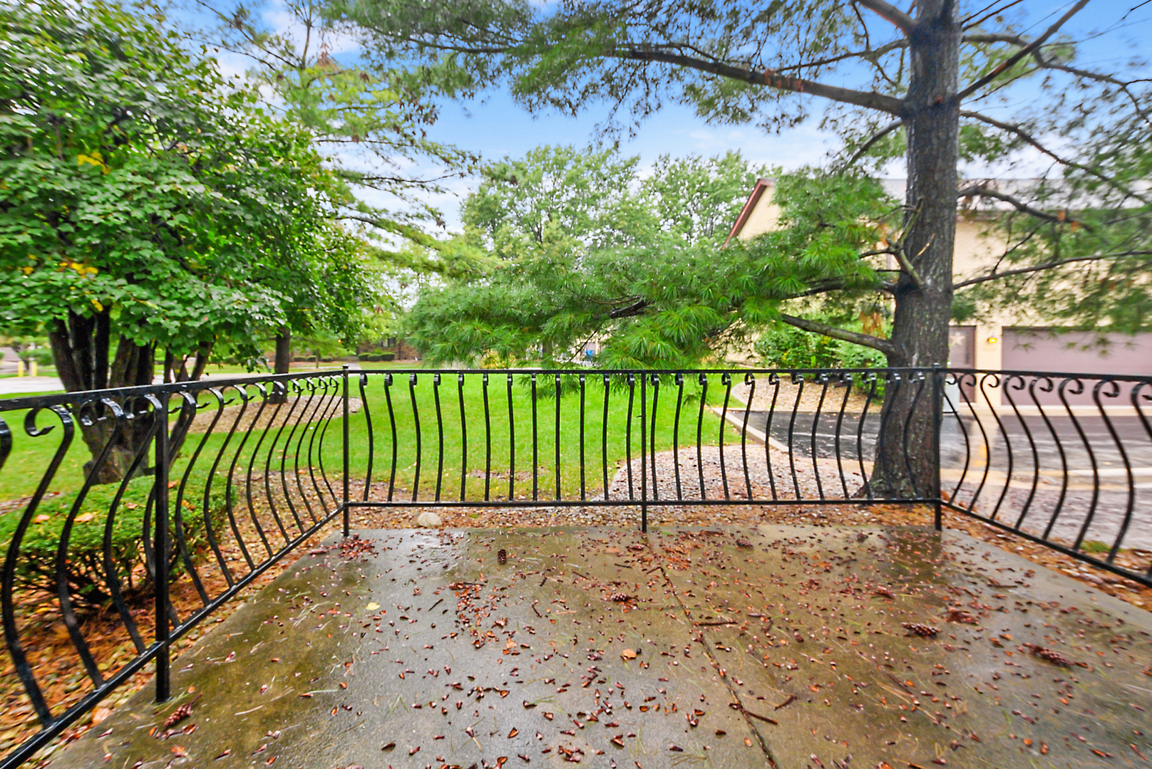 Concrete patio with decorative black railing beneath mature trees, overlooking a green lawn.