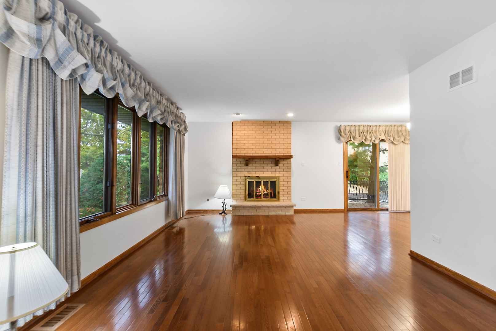 Living room with a brick fireplace, polished hardwood floors, and large windows overlooking greenery.