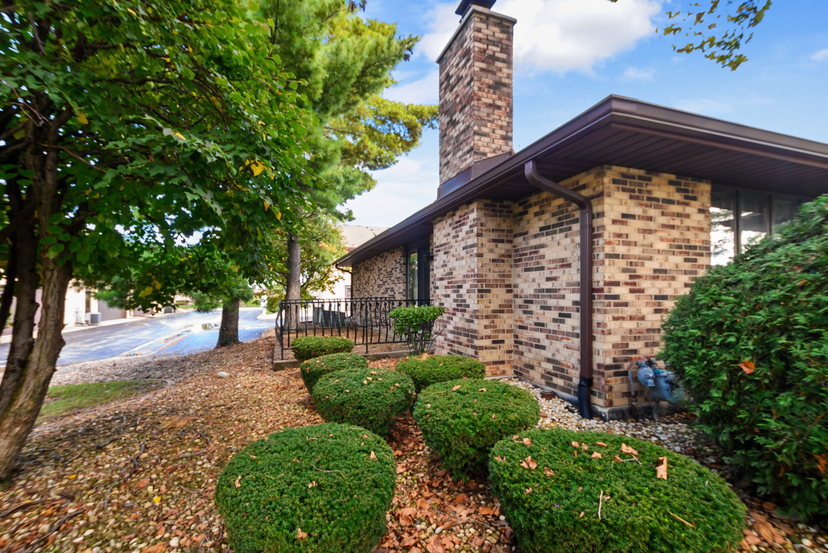 Brick home's exterior with chimney, black railing, and trimmed shrubs beneath mature trees.