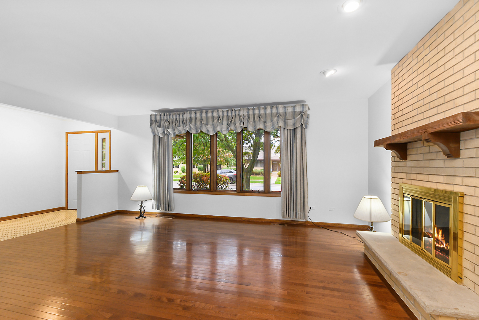 Empty living room with hardwood floors, a large curtained window, and a brick fireplace.