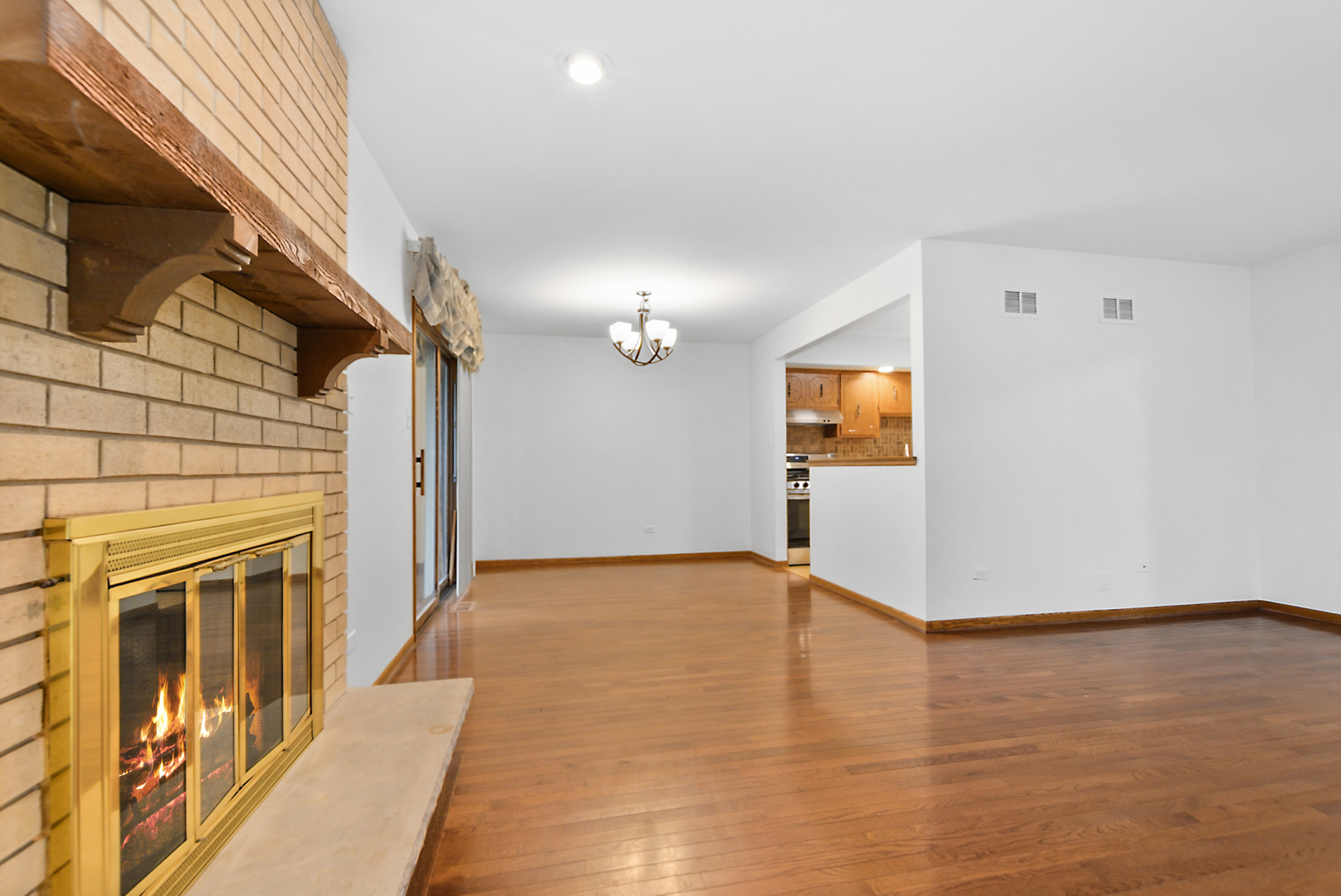 Empty living and dining area with hardwood floors and a brick fireplace with brass surround.