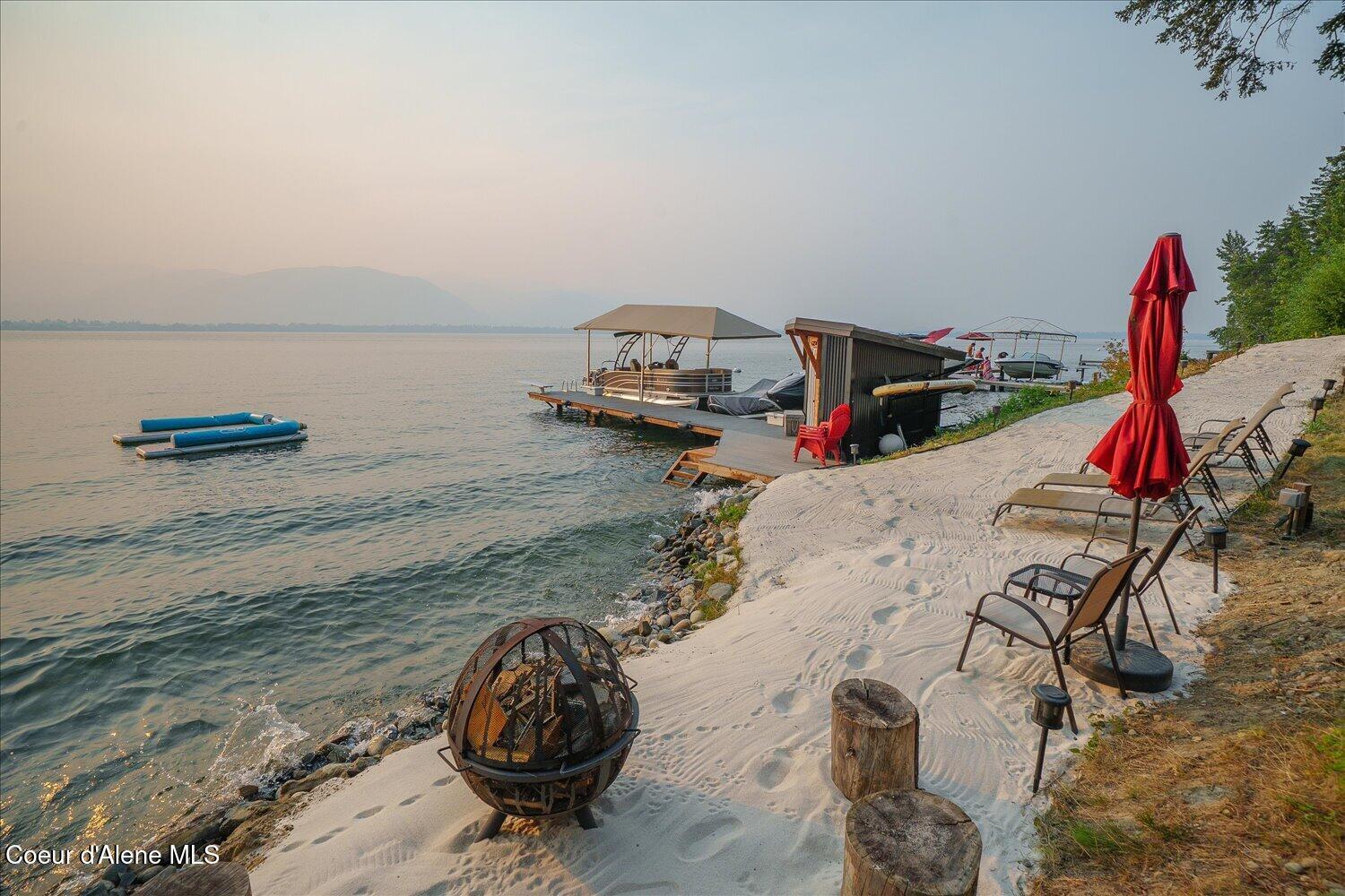 Sandy lakeshore with docked boats, lounge chairs, and a hazy mountain view across the water.