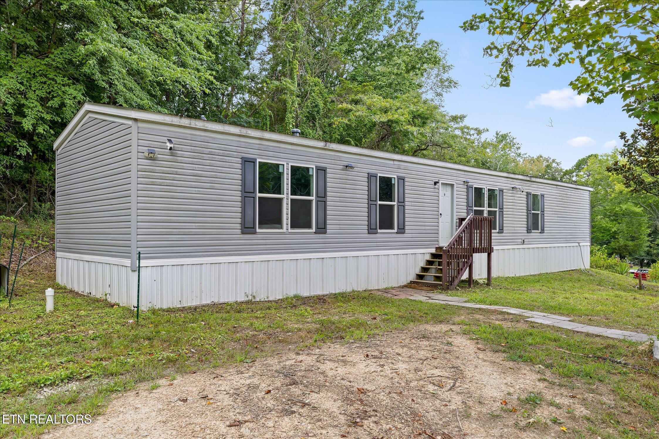 Gray manufactured home with dark shutters and wooden entry steps on a wooded lot.