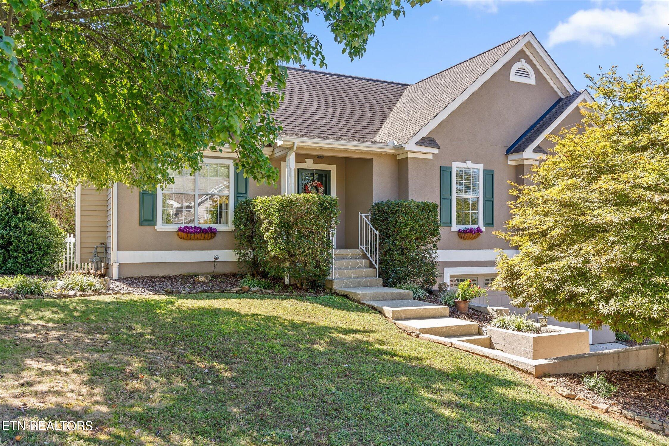 Tan house with green shutters, front steps, and a landscaped lawn shaded by mature trees.