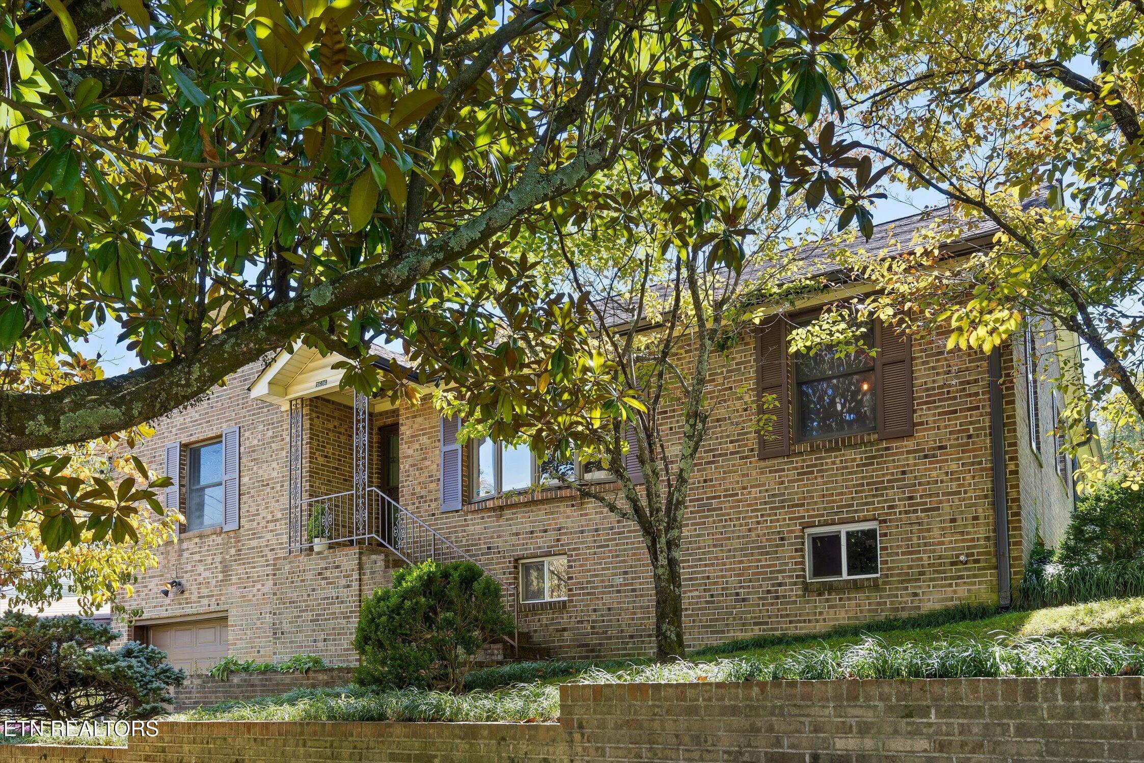 Two-story brick house partly shaded by mature trees, with a covered entry and landscaped front yard.