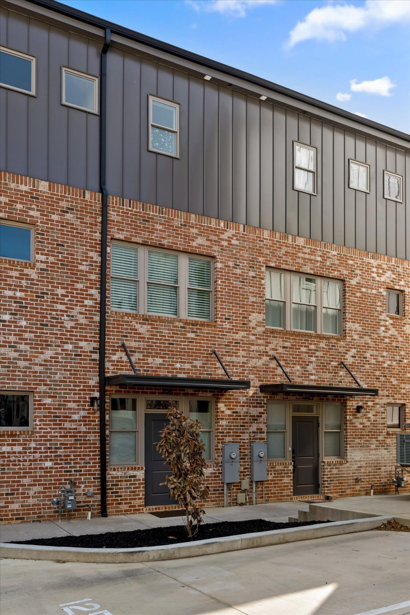 Attached brick townhomes with gray siding, black entry awnings, and a young tree in front