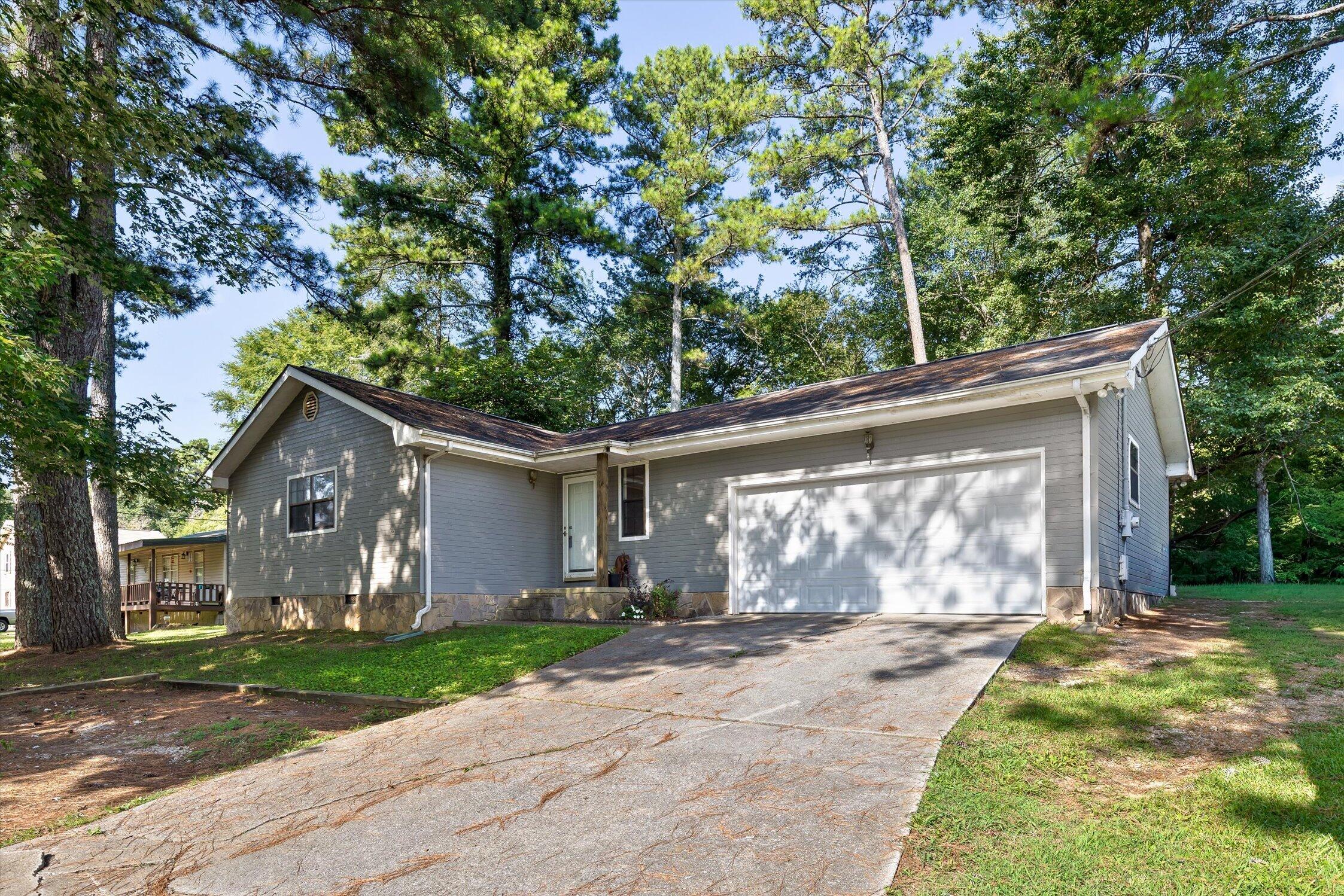 Gray single-story home with an attached two-car garage, stone foundation, and mature trees.