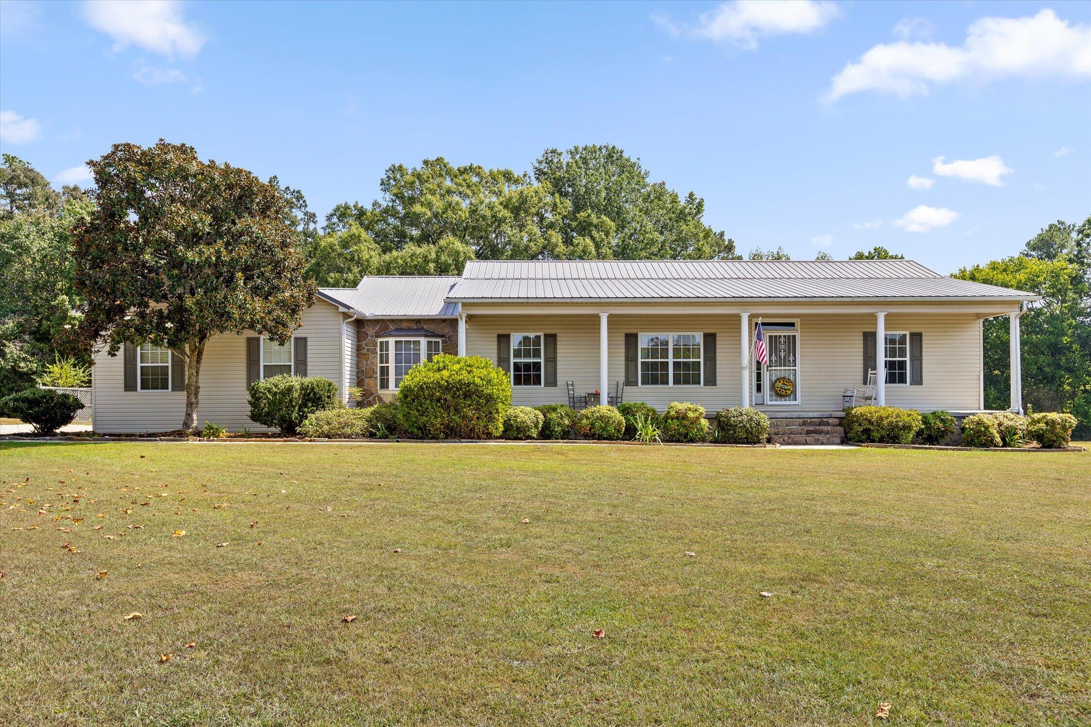 Single-story beige home with a long covered porch, white metal roof, stone bay-window accent, and broad front lawn.