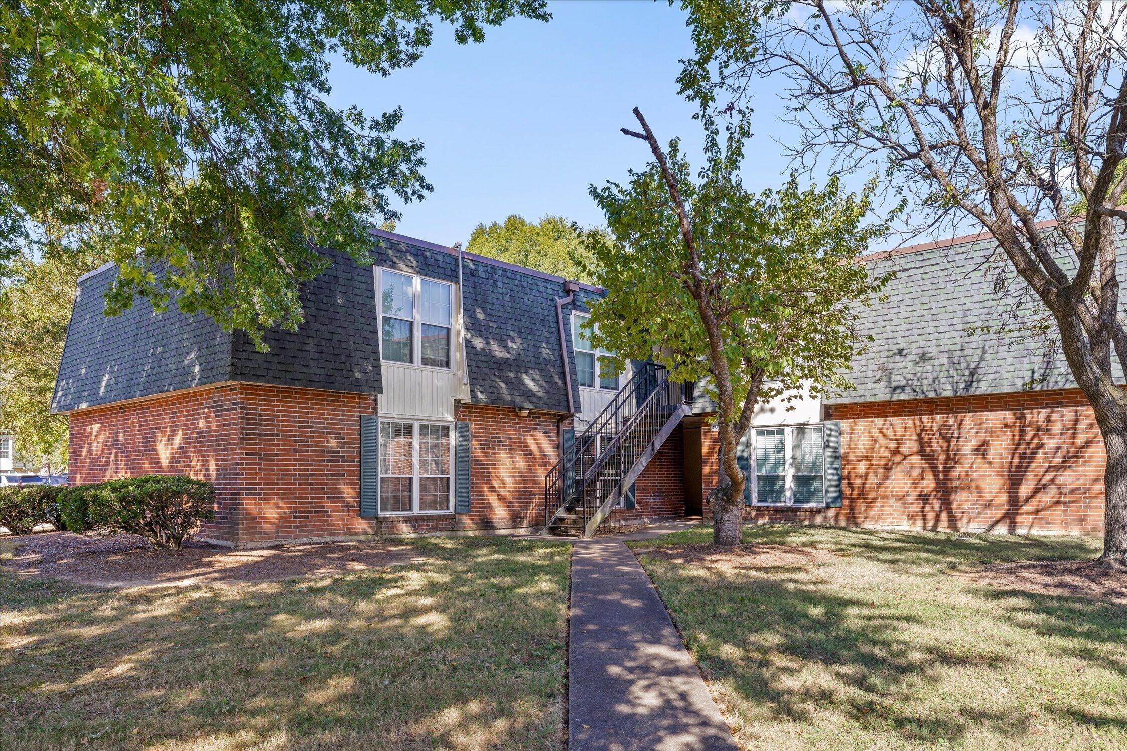 Two-story brick residential building with a dark shingle roof and exterior staircase beside a tree-lined walkway.