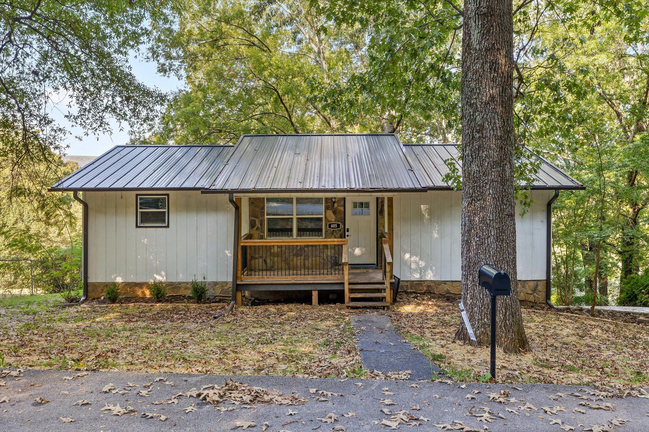 White-sided house with a metal roof, covered porch, and front steps beneath mature trees.