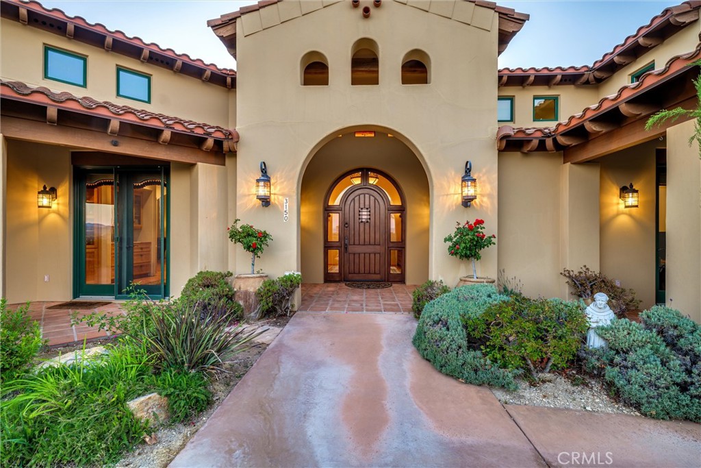 Stucco home entrance with an arched wooden door, terracotta roof, and landscaped front walkway.