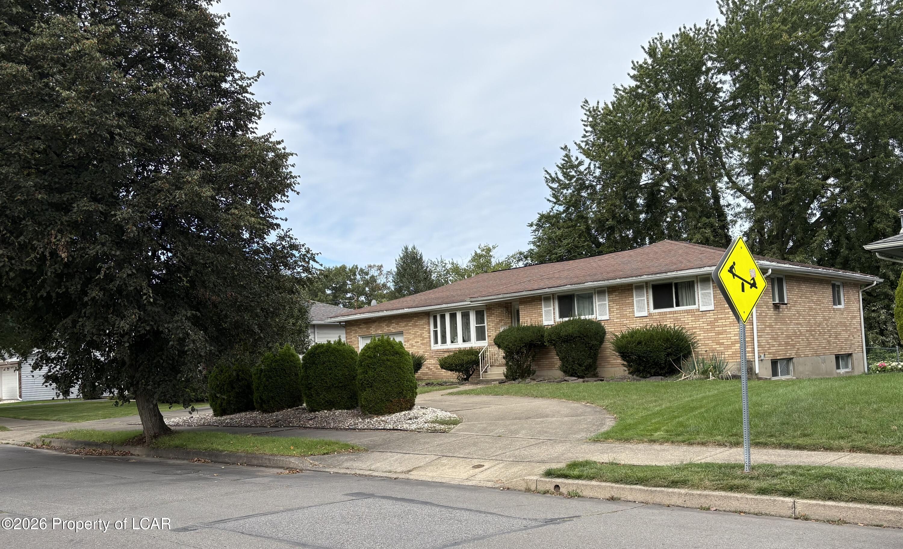 Single-story brick ranch with clipped shrubs, broad lawn, and a yellow crossing sign in the foreground.