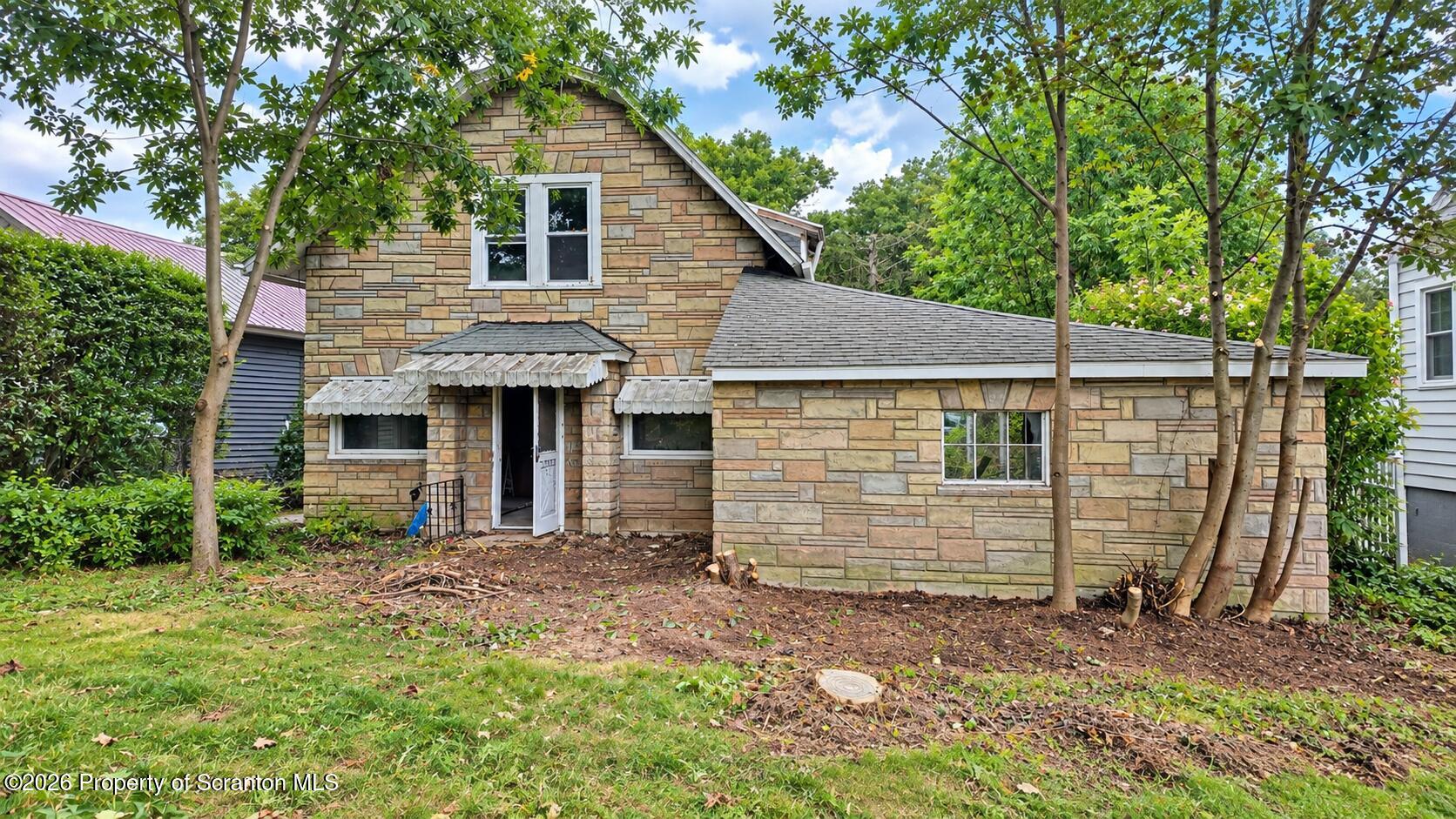 Stone-faced house with a steep gable and open front door, framed by mature trees.