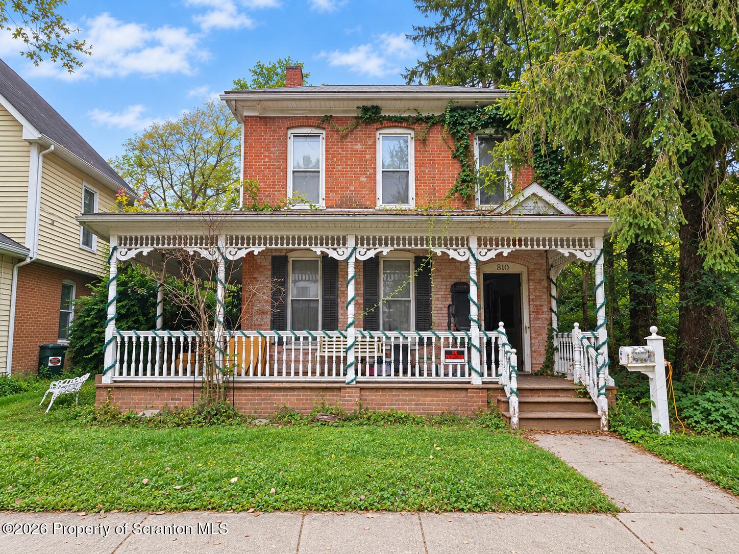 Two-story red-brick house with an ornate white porch, leafy trees, and a front lawn.