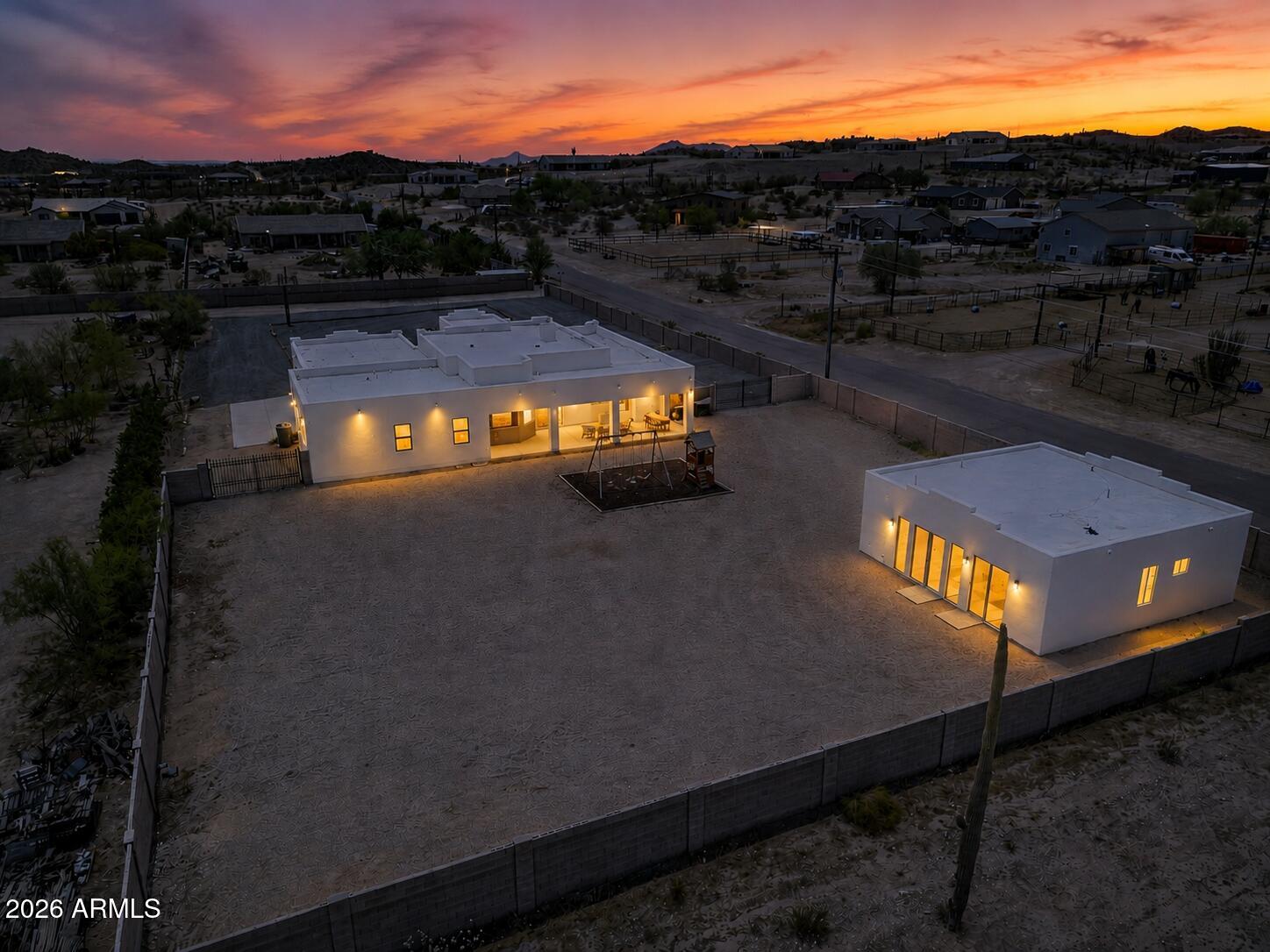 Aerial view of two illuminated white buildings in an enclosed gravel lot beneath a vivid sunset.