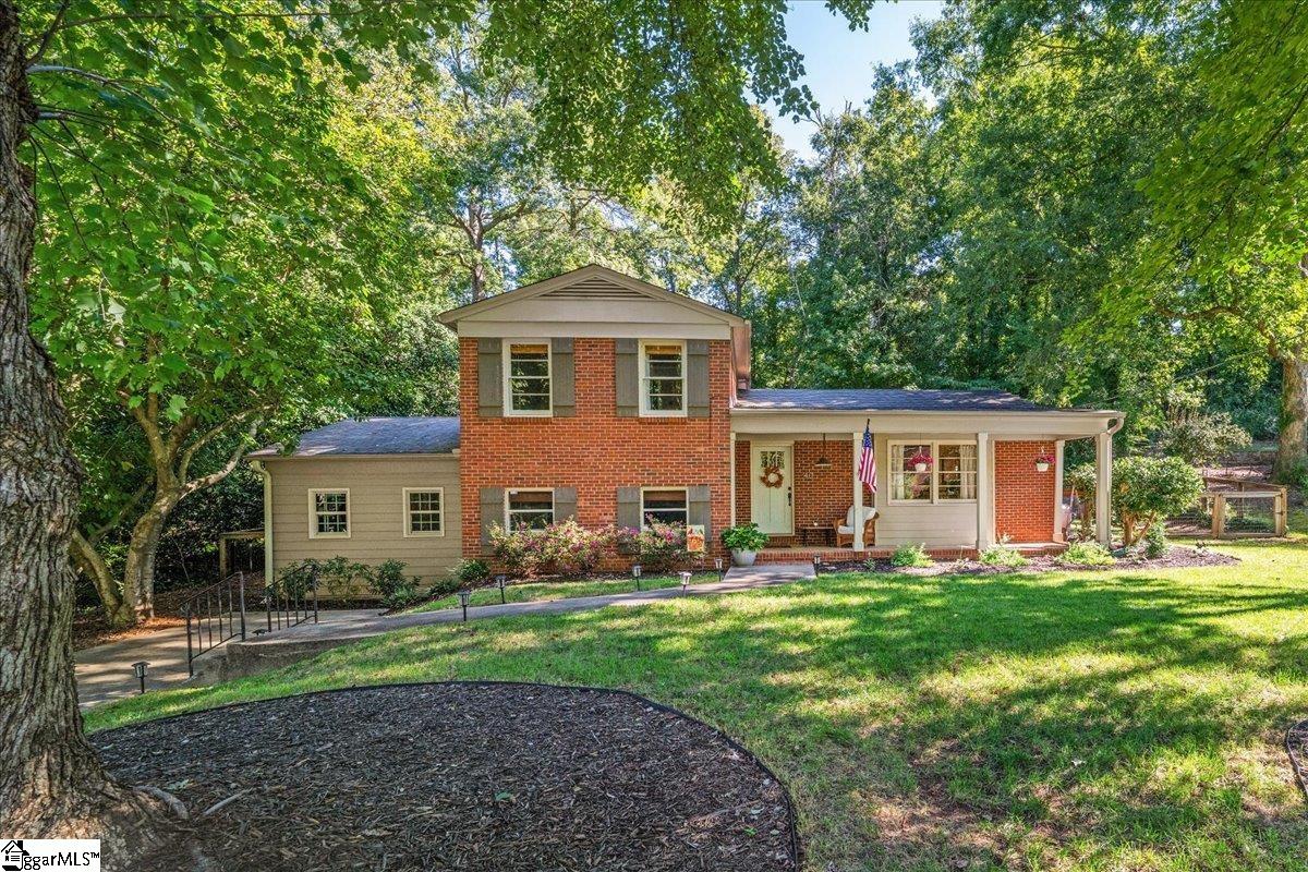 Two-story brick and beige home with a covered porch, flower beds, and a lawn beneath mature trees.