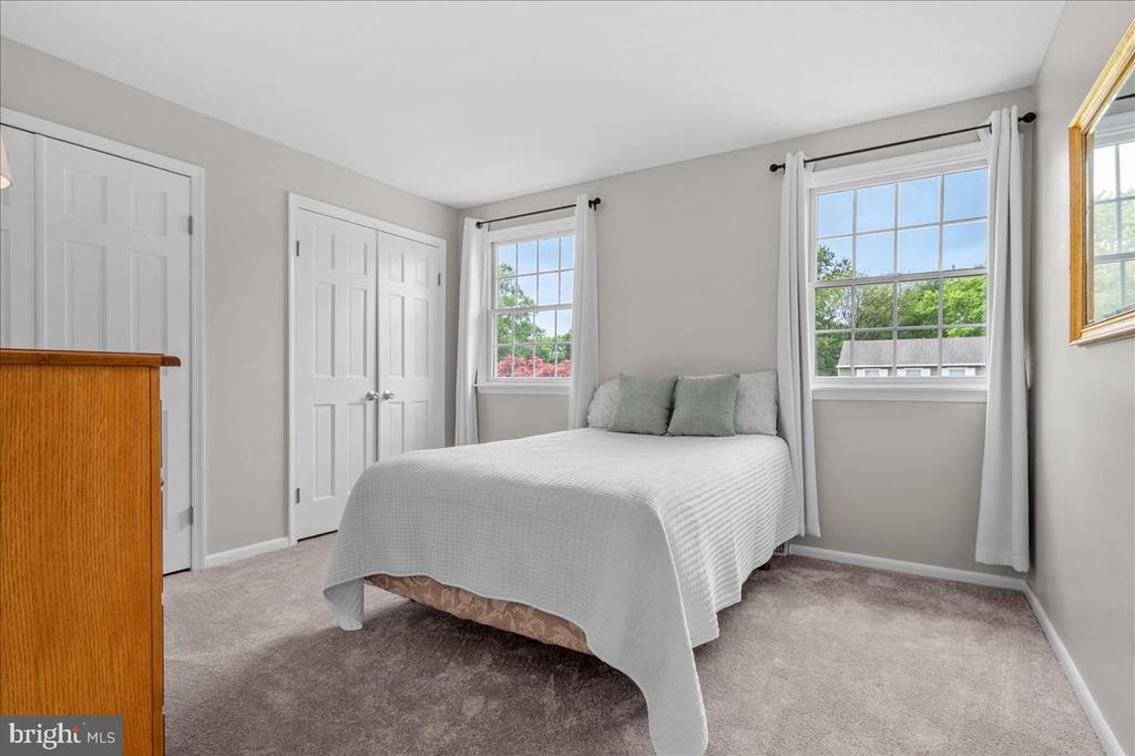 Bedroom with a white-covered bed, beige carpet, double closet doors, and two windows overlooking greenery.