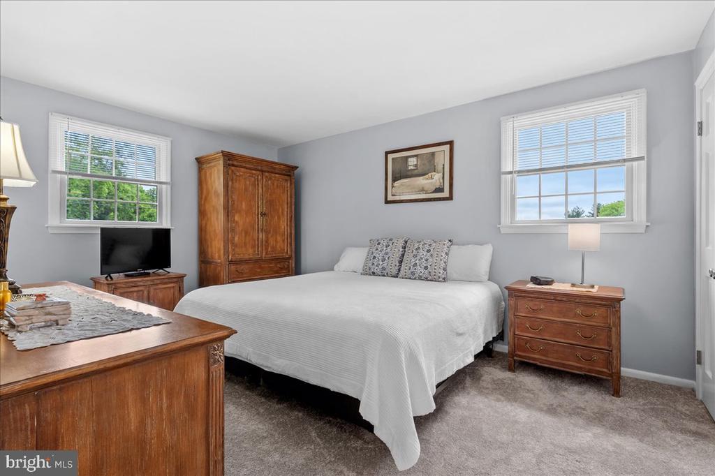 Bedroom with a white bed, dark wood armoire, and two windows with blinds.