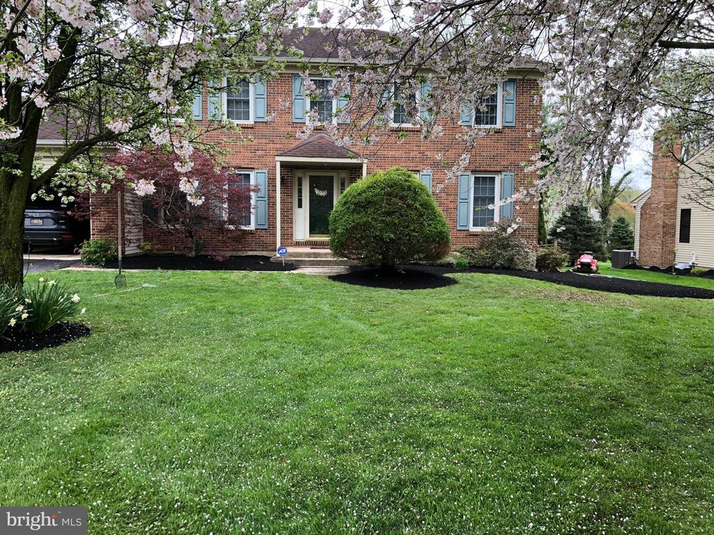 Two-story brick house with blue shutters and a covered entry, framed by flowering trees and a green lawn.