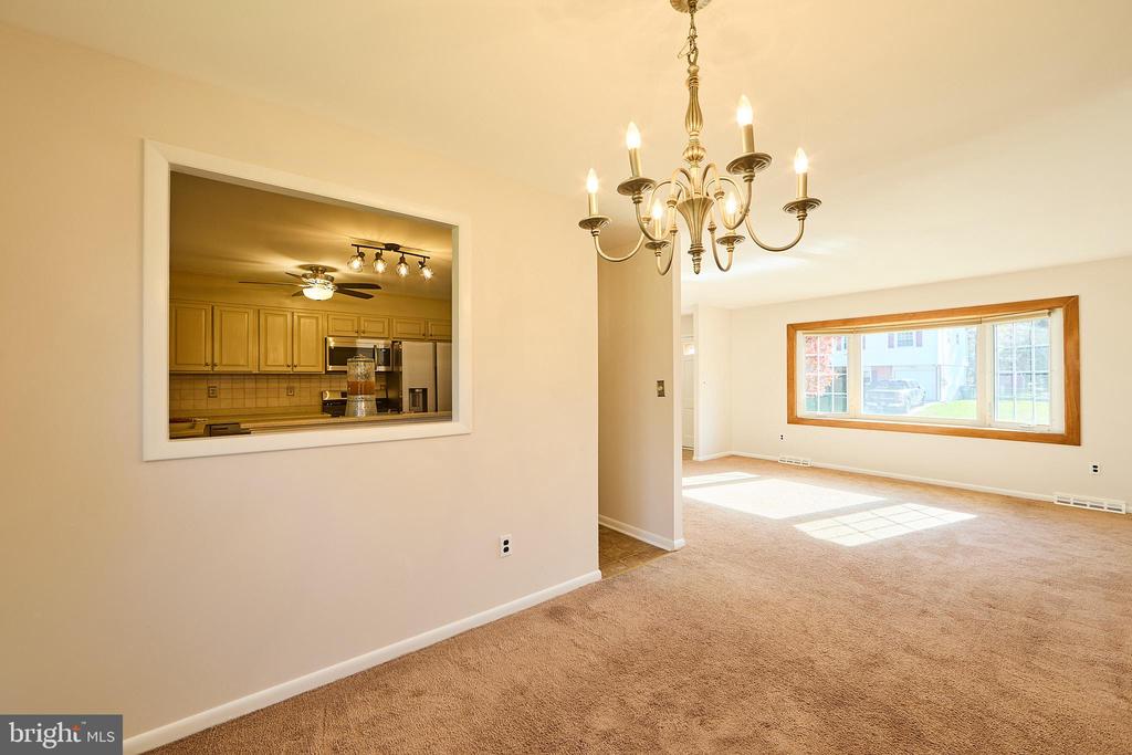 Empty living room with a brass chandelier, carpeted floor, and large picture window beside a kitchen pass-through.
