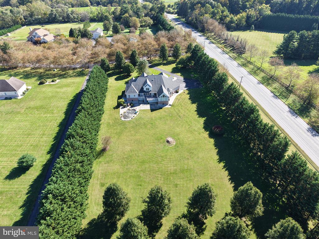 Aerial view of a home with a broad lawn, evergreen tree rows, and a road alongside the property.