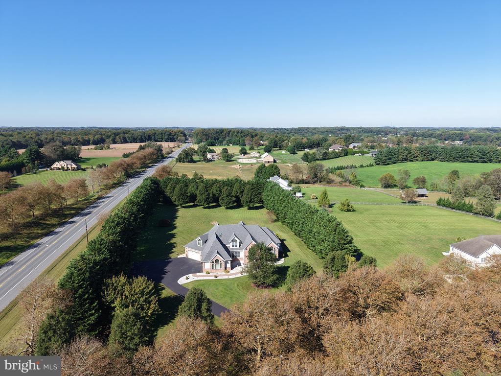 Aerial view of a detached house with a curved driveway, evergreen rows, and open fields beside a road.