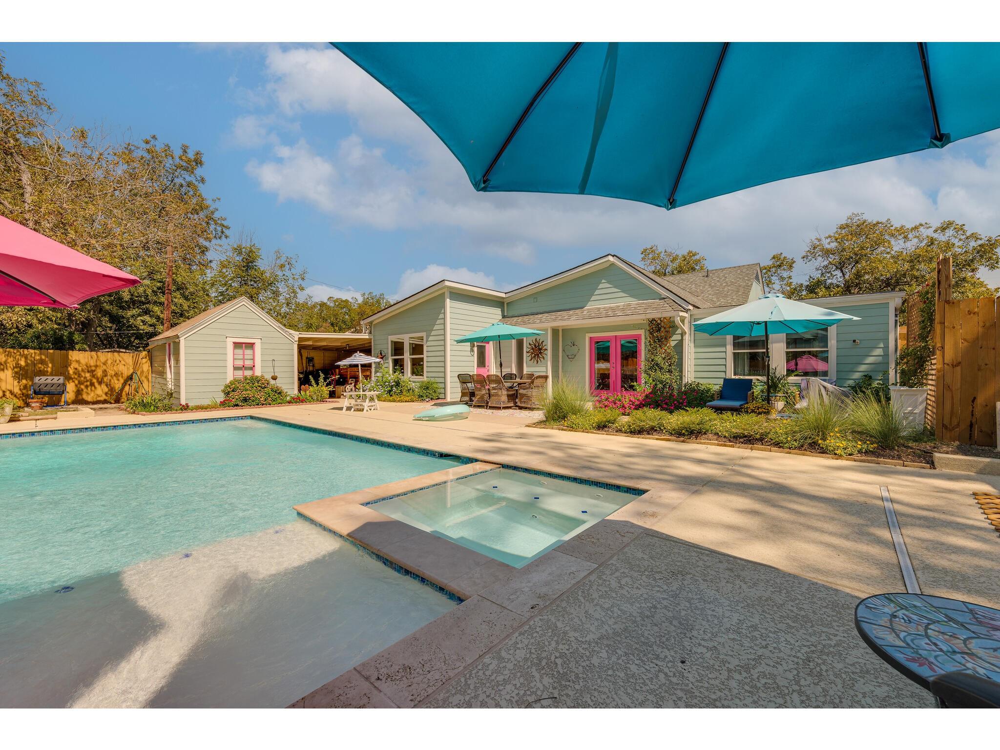 Backyard swimming pool and spa beside a pale green home, shaded by colorful umbrellas.