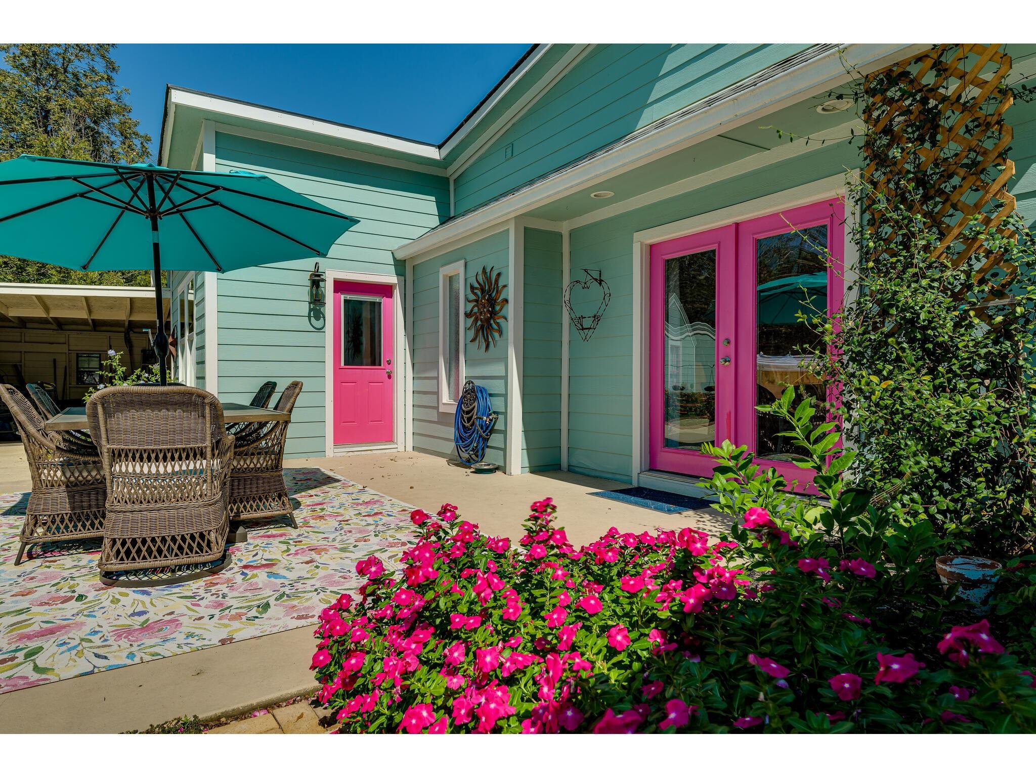 Patio with aqua siding, bright pink doors, wicker dining set, teal umbrella, and flowering shrubs.