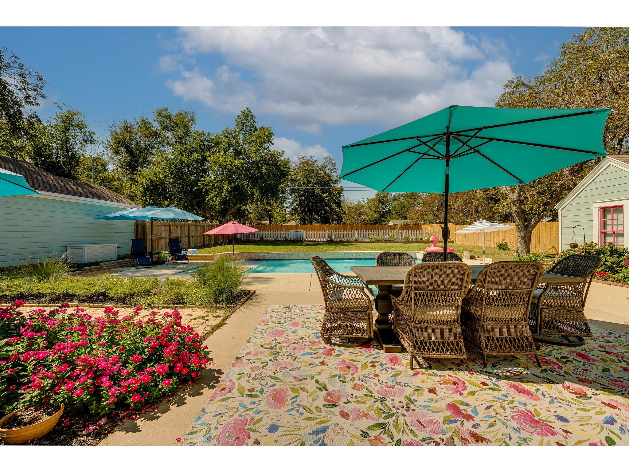 Backyard pool beside a patio dining set under a large turquoise umbrella and surrounded by flowering beds.