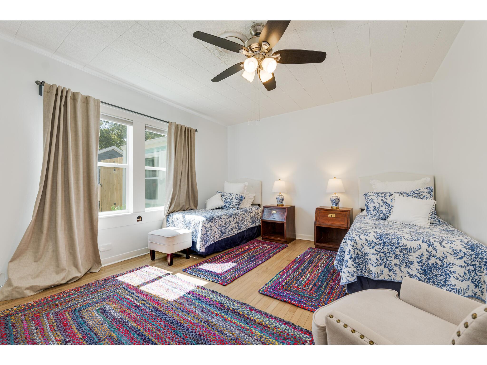 Bedroom with two floral-covered beds, colorful braided rugs, and beige curtains.