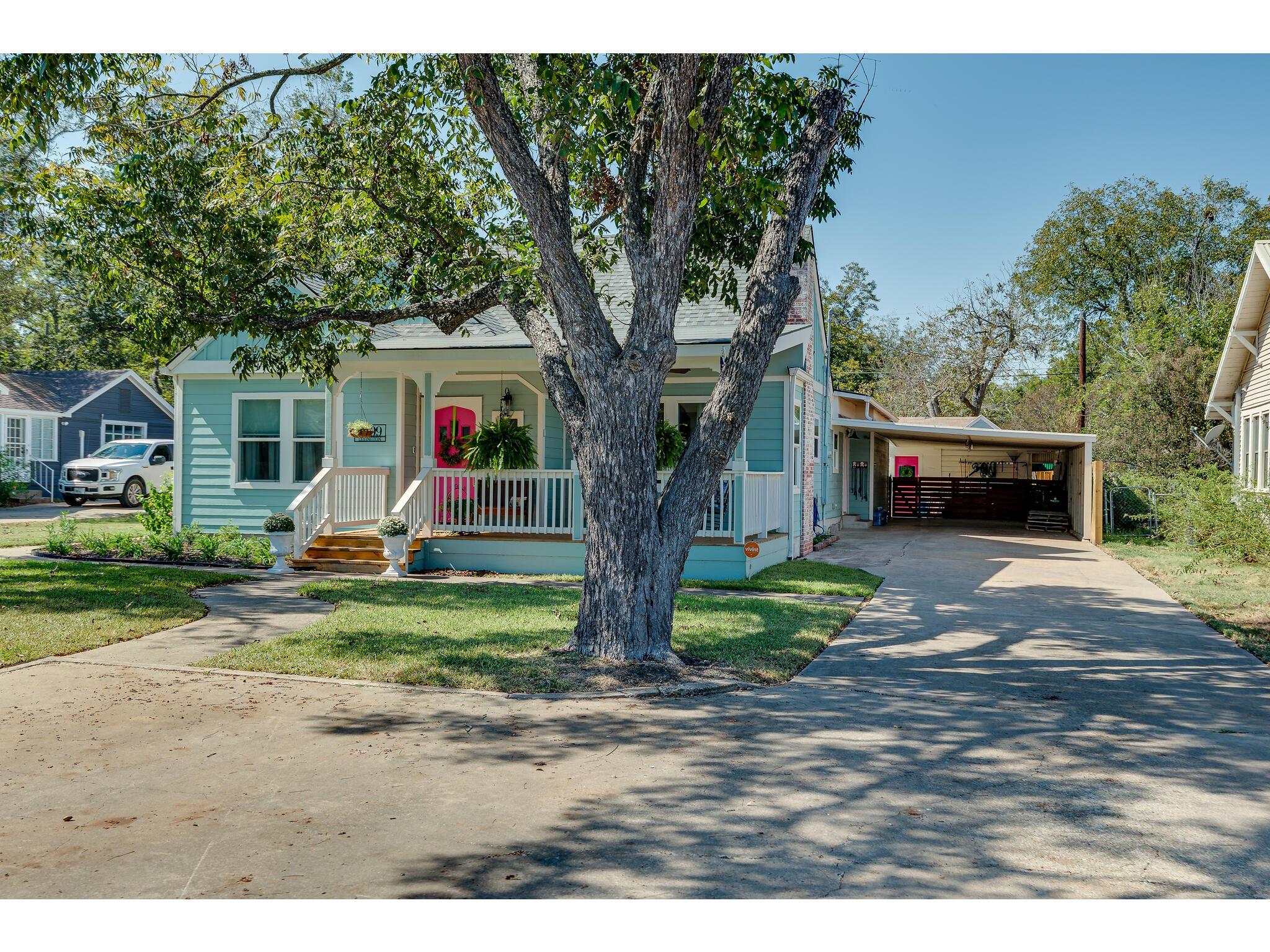 Light blue house with a white porch and pink door, partly obscured by a large tree beside the driveway.