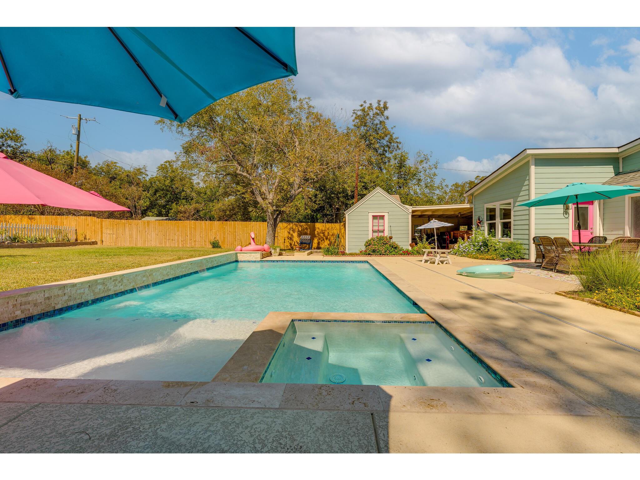 Backyard pool and spa beside a pale green home, with colorful umbrellas and patio seating.