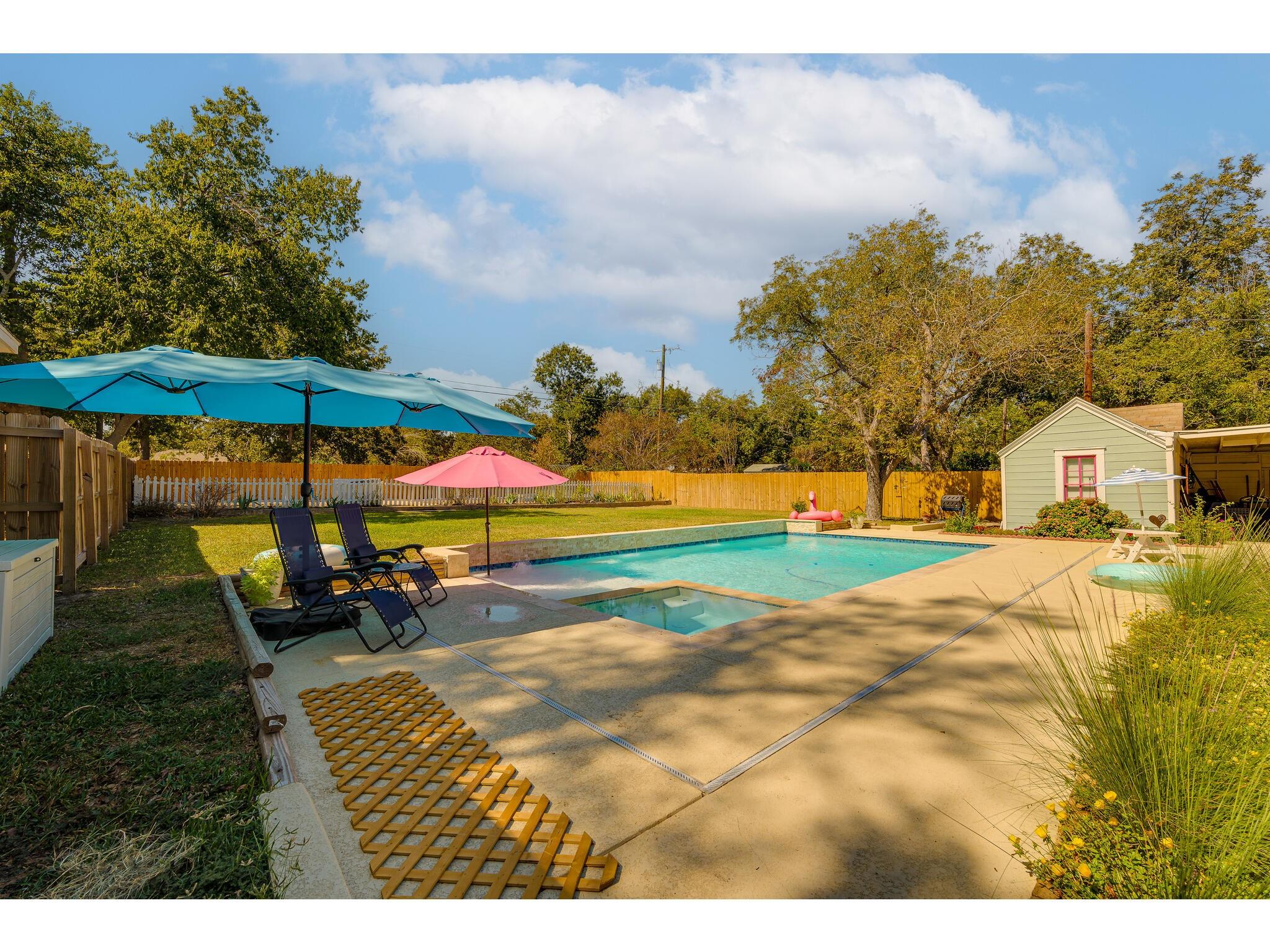 Backyard pool and patio with colorful umbrellas, lounge chairs, fenced lawn, and a small shed.