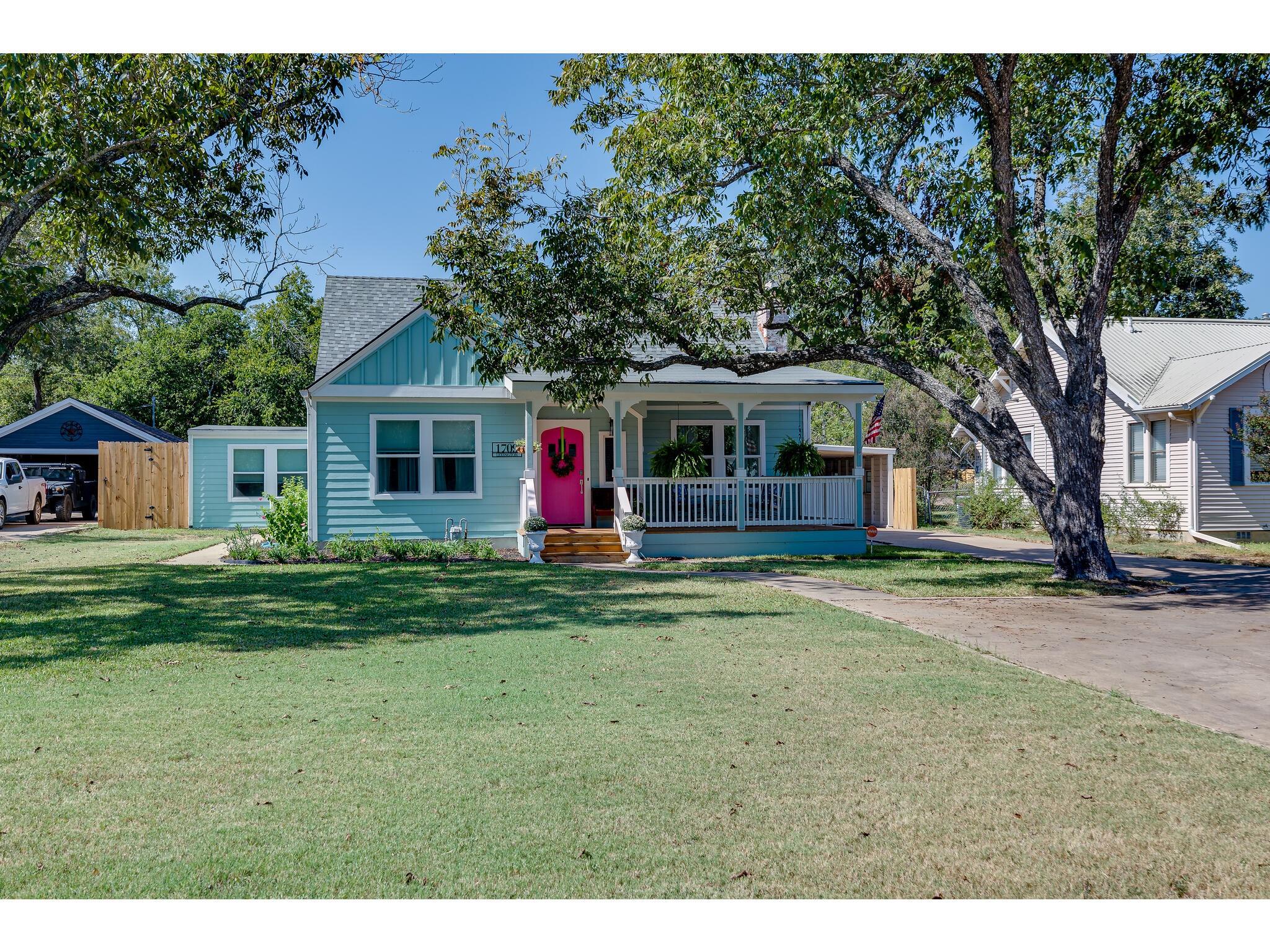 Light blue single-story house with a covered porch, bright pink front door, and large shade tree.