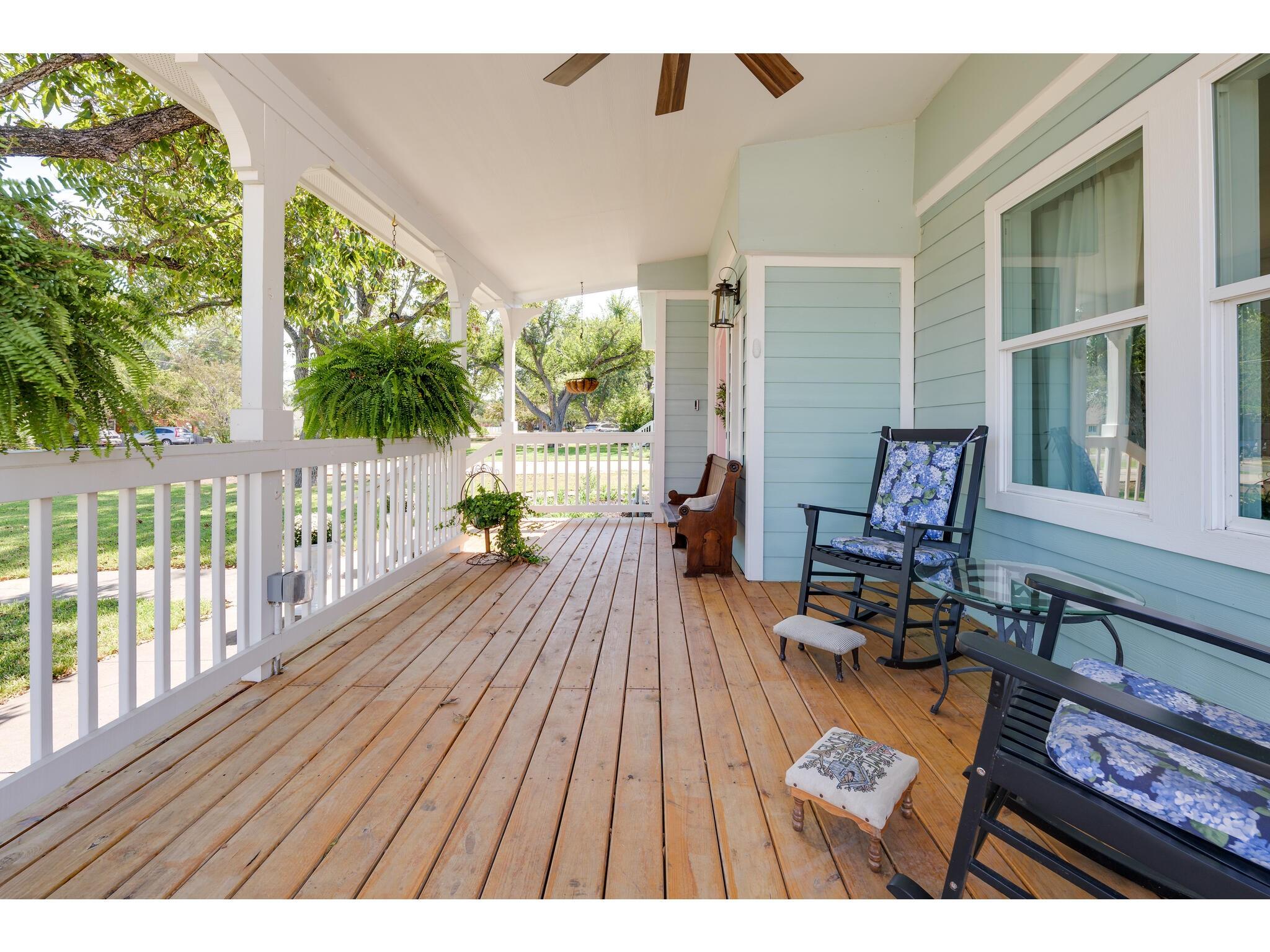 Covered wooden porch with white railings, light blue siding, hanging ferns, and floral-cushioned chairs.