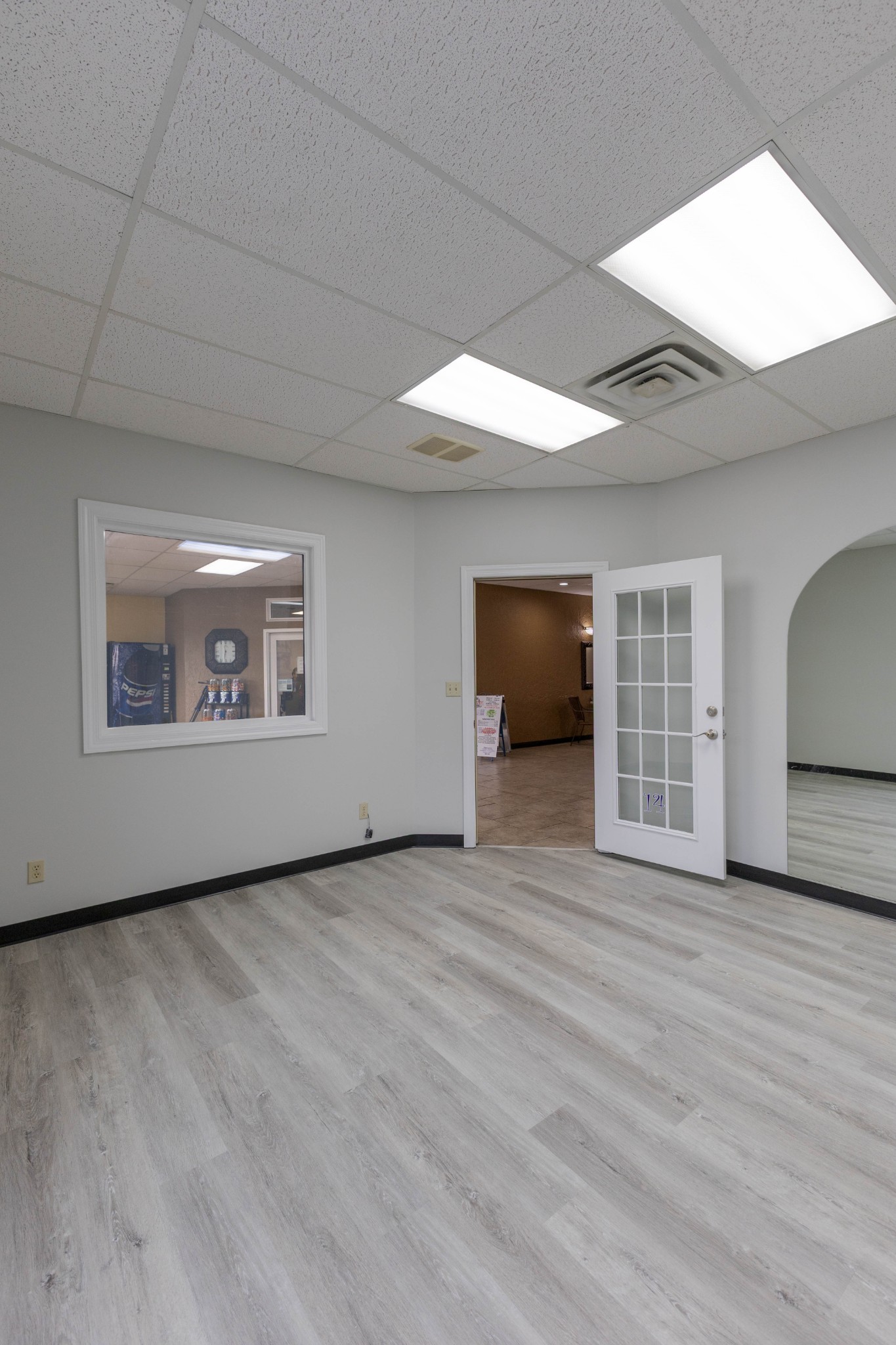 Vacant commercial interior with gray plank flooring, a glass-paneled door, and a window overlooking a Pepsi vending machine.