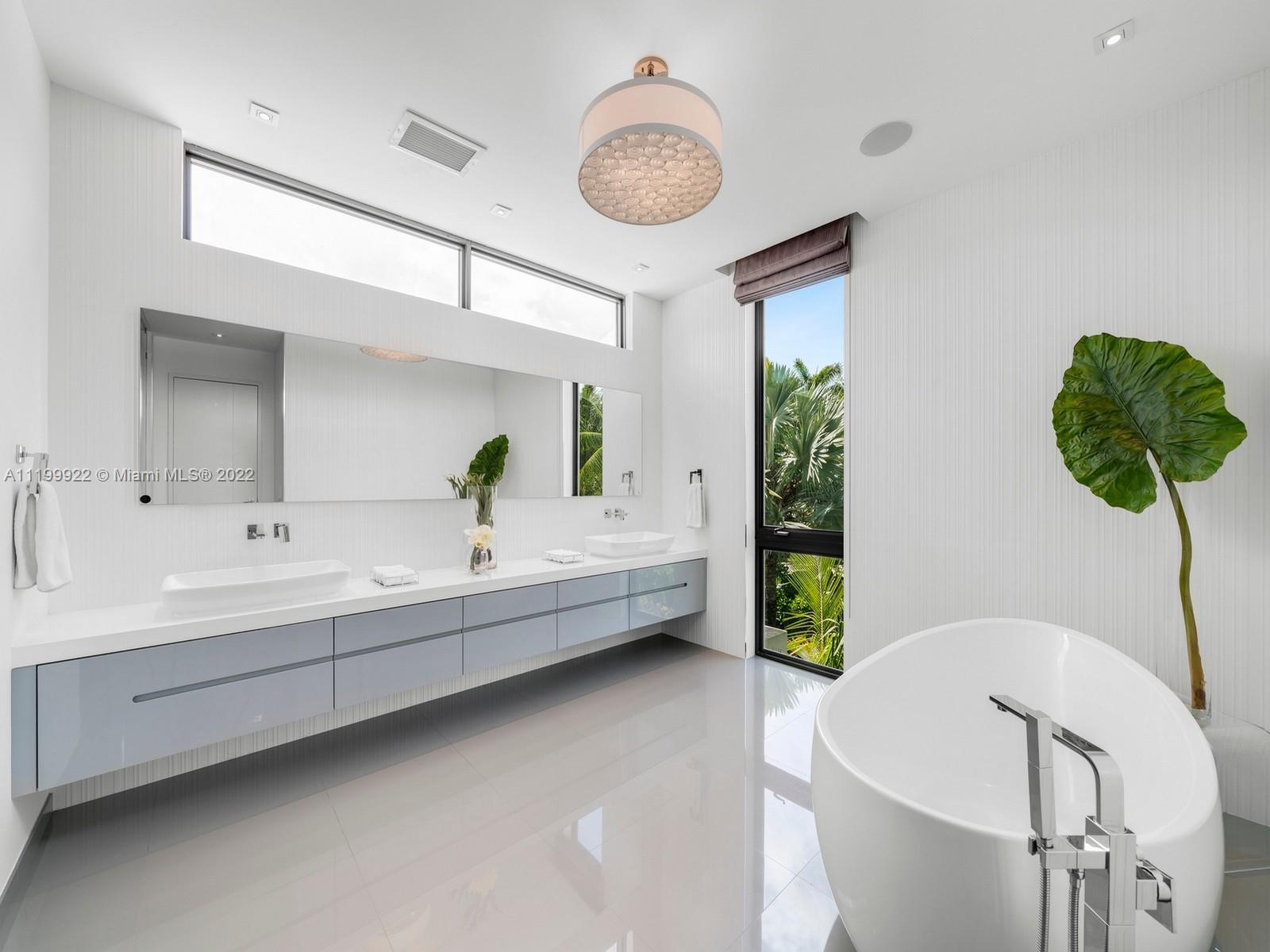 Modern bathroom with a freestanding soaking tub, double vanity, and tall windows overlooking tropical greenery.