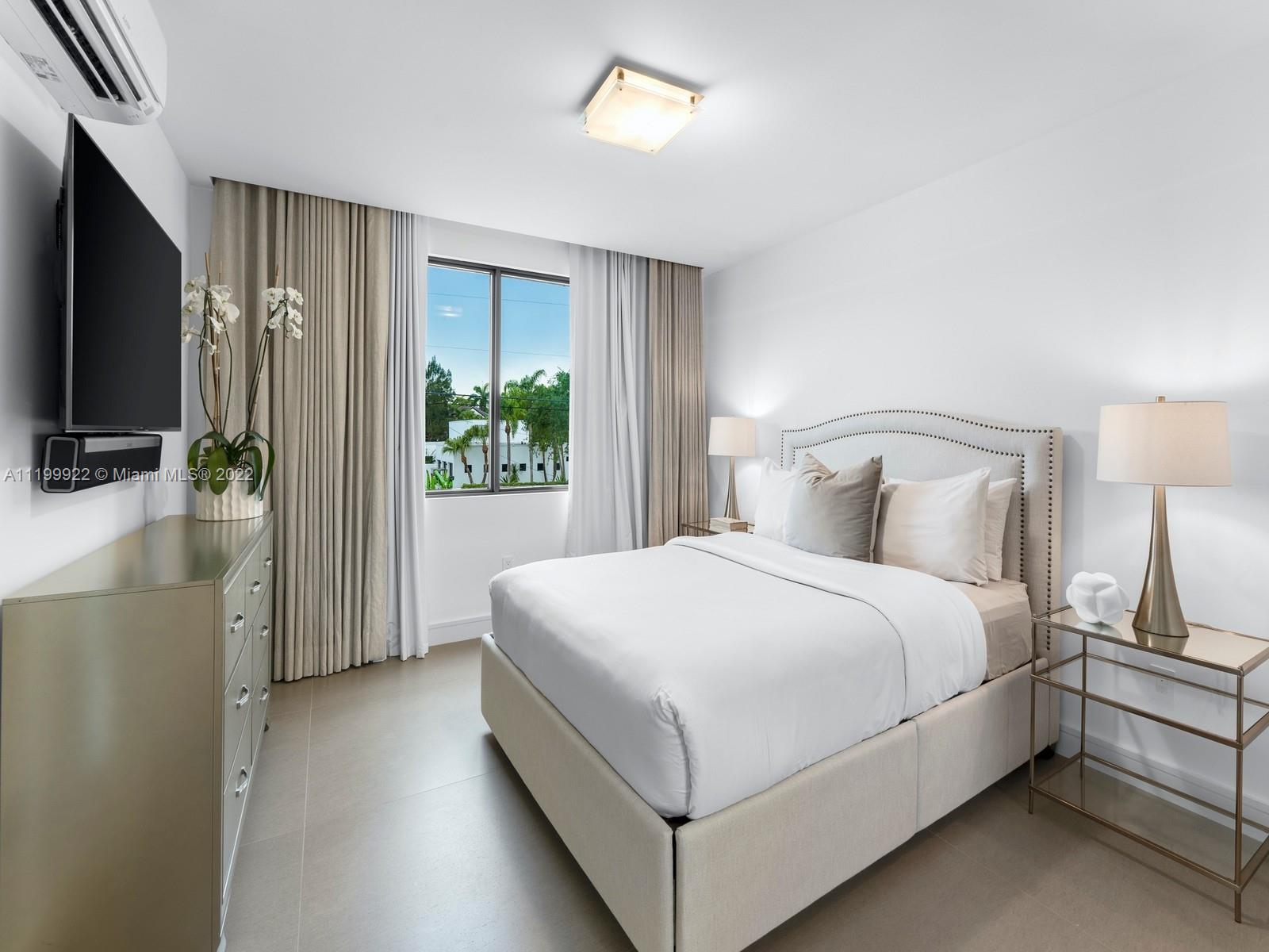 Staged bedroom with a white bed, curved nailhead-trim headboard, beige curtains, and a window.