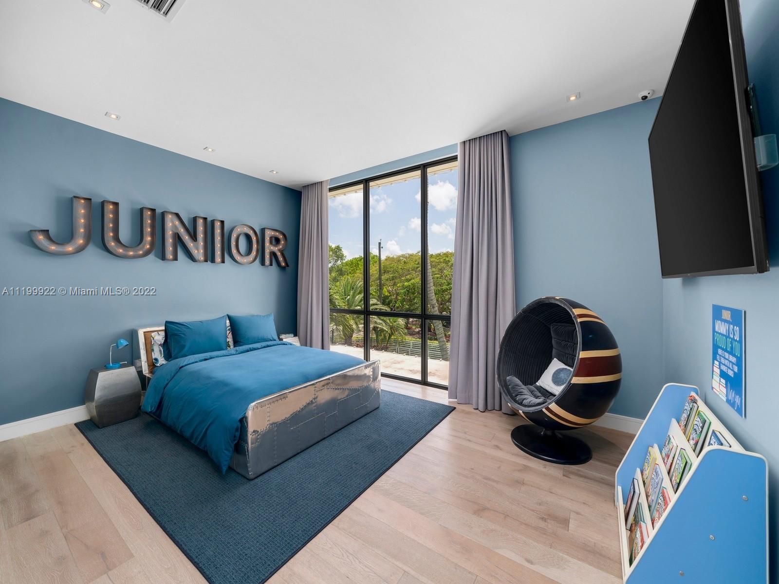 Blue bedroom with a teal bed, illuminated JUNIOR letters, round chair, and tall windows overlooking trees.