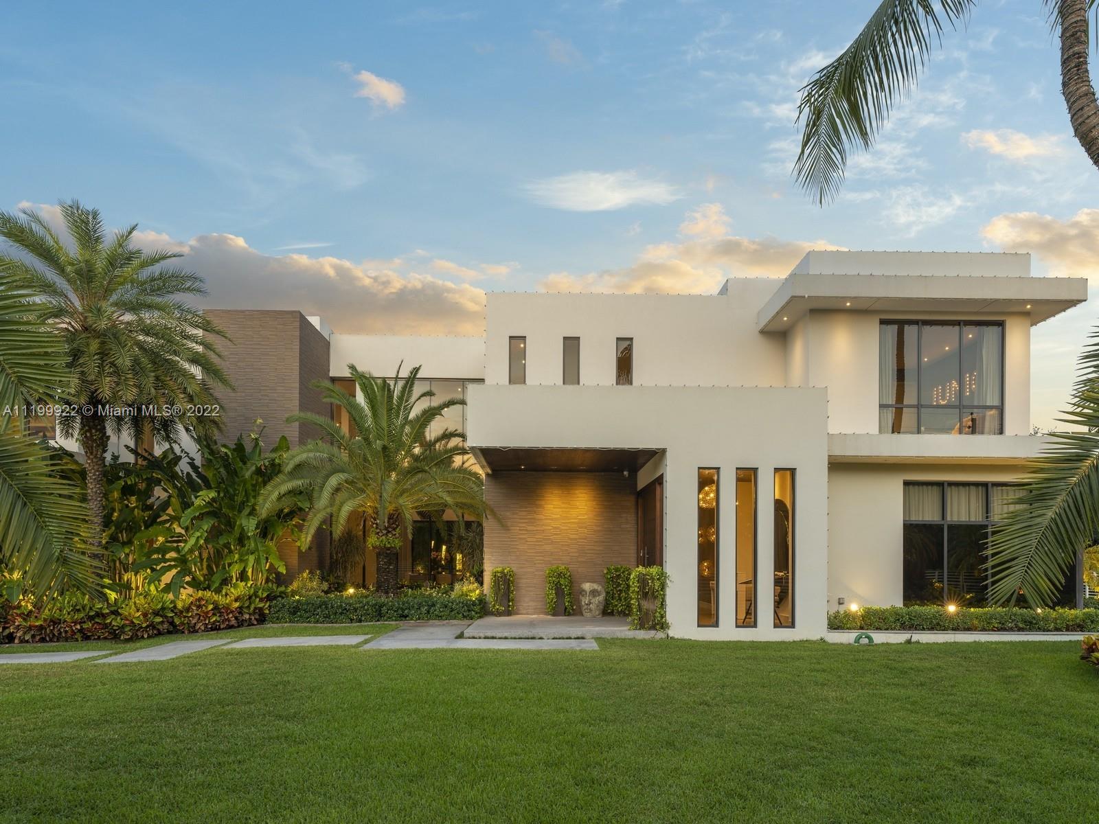 Modern white home with tall windows, tropical landscaping, and a broad front lawn at dusk.